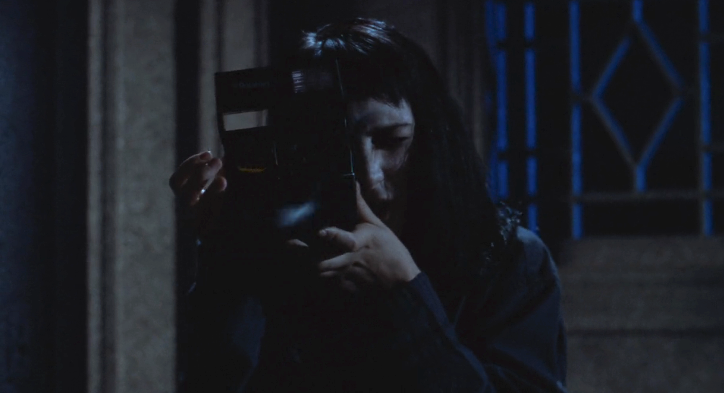 Winona Ryder is holding a Polaroid camera up to their face, seemingly taking a picture. The setting appears dark and moody, suggesting a suspenseful or eerie atmosphere