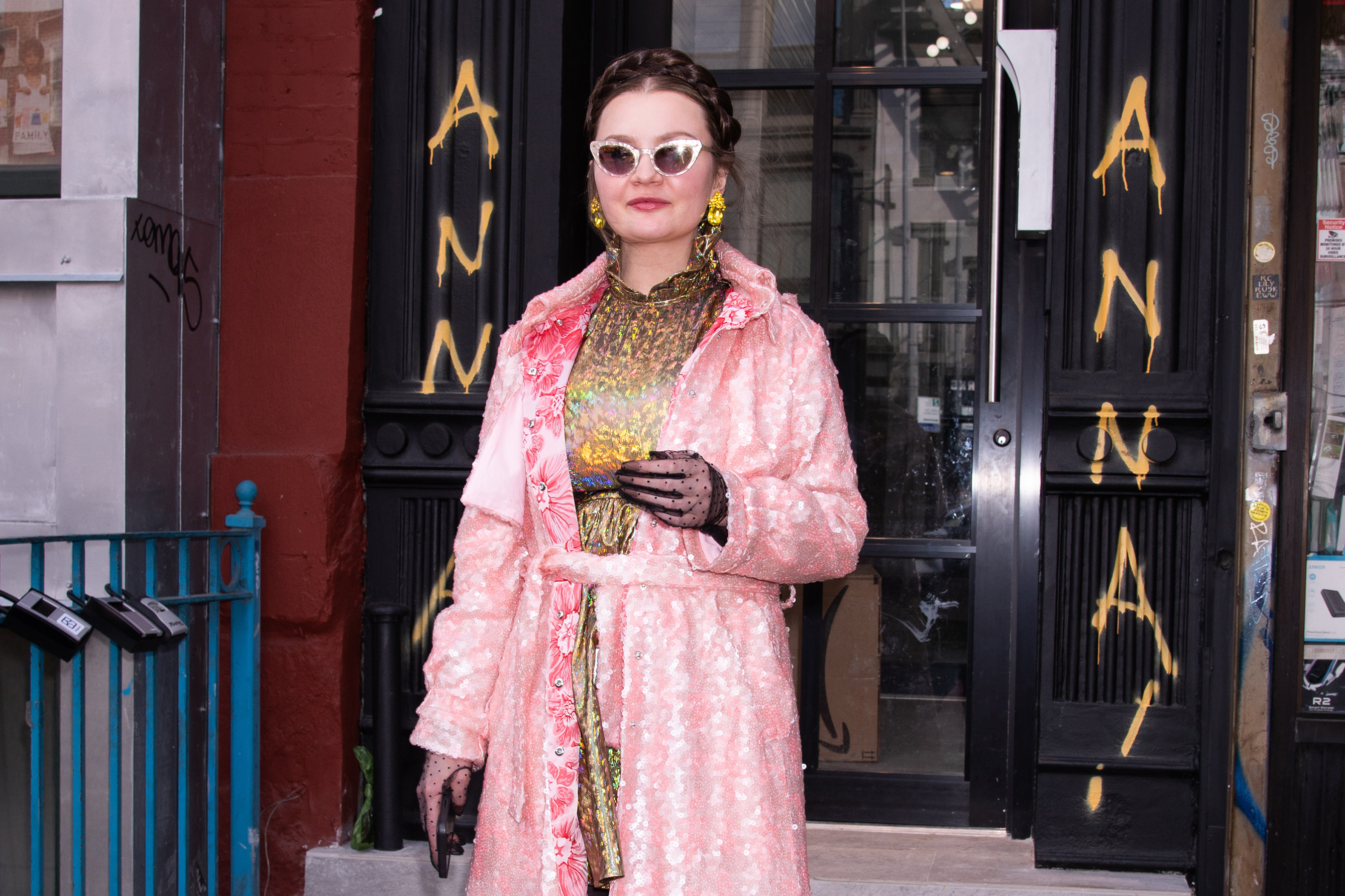 Anna Sorokin stands in front of a storefront wearing a pink textured coat, patterned dress, gloves, and sunglasses, with her hair in braids