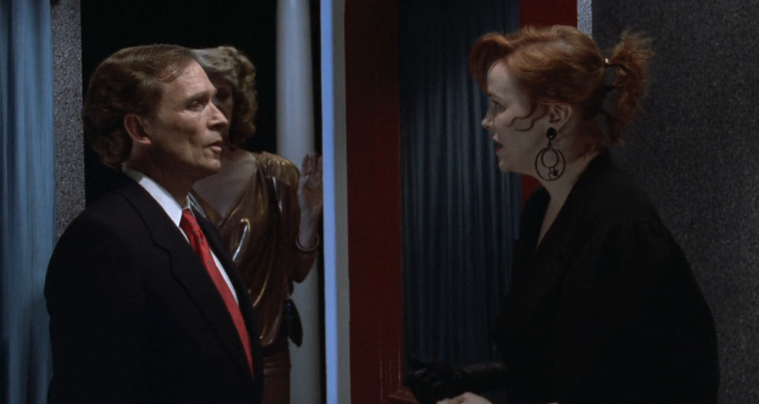 Dick Cavett speaks with Catherine O'Hara, who wears a dark outfit with large earrings. A person in a shiny dress is in the background