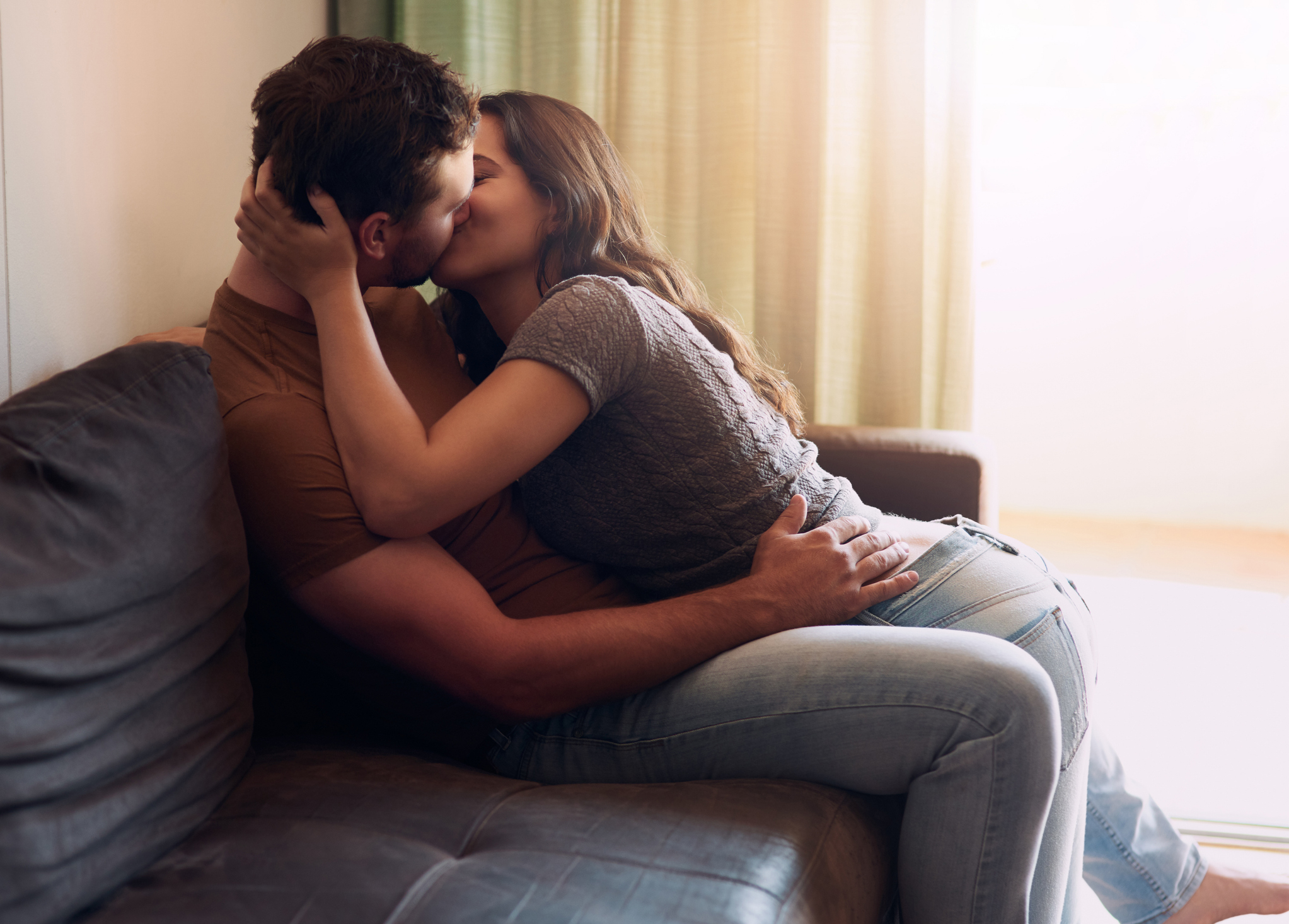 A couple is passionately kissing on a couch, with the woman sitting on the man's lap and their arms wrapped around each other