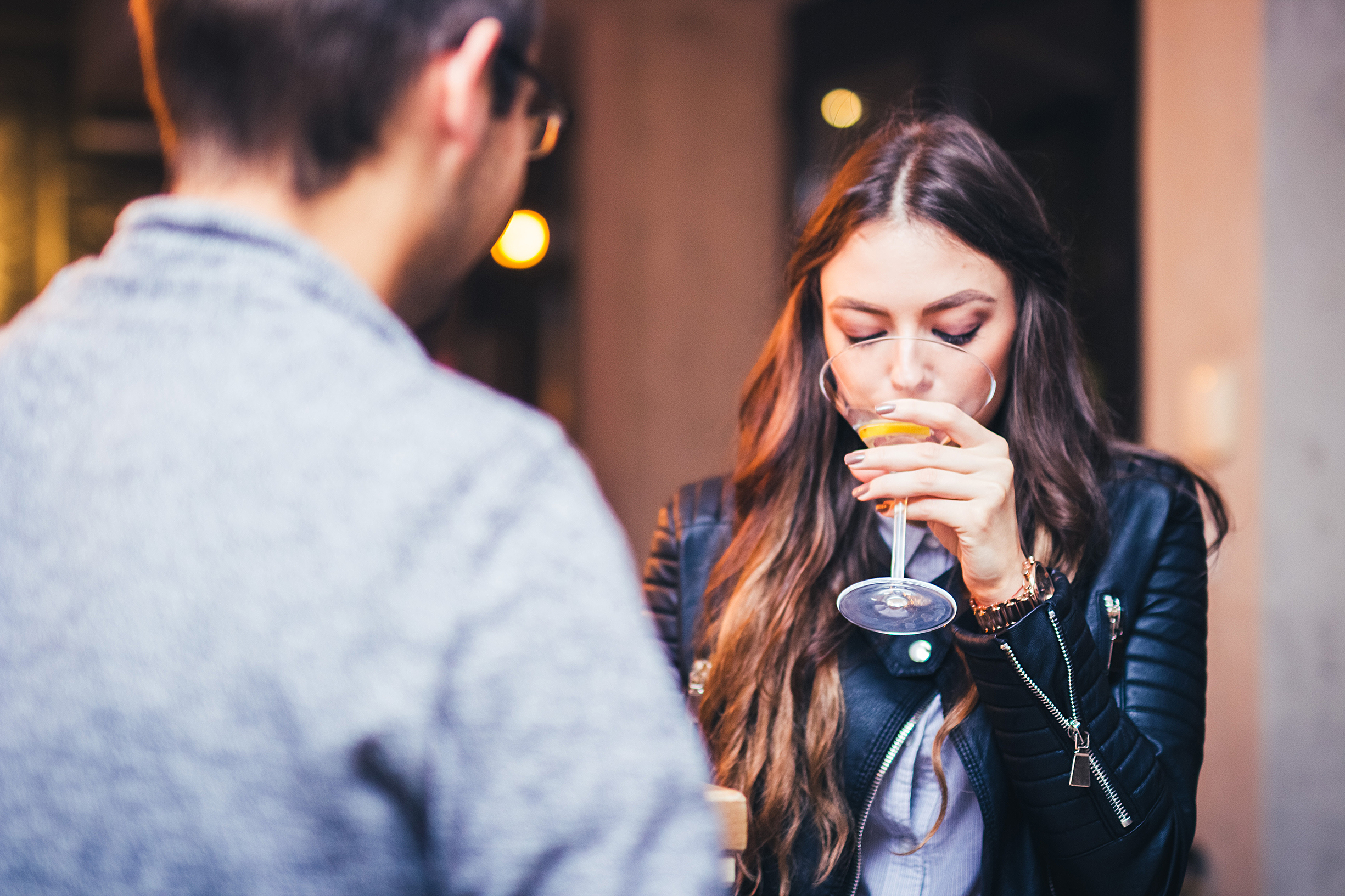 A woman with long hair and a leather jacket sips a drink while sitting across a man in a casual sweater