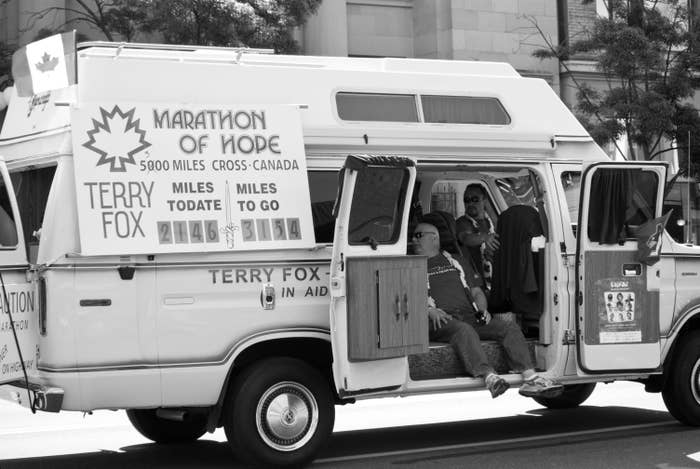 Terry Fox sitting in a van labeled "Marathon of Hope" with signs indicating a 5000-mile run across Canada. Other unidentified people are also present in the van