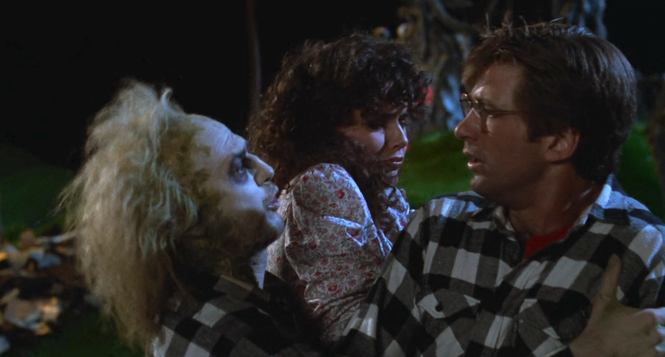 Michael Keaton as Beetlejuice grips Geena Davis and Alec Baldwin, who are looking at each other. Geena is wearing a floral dress, and Alec is in a plaid shirt