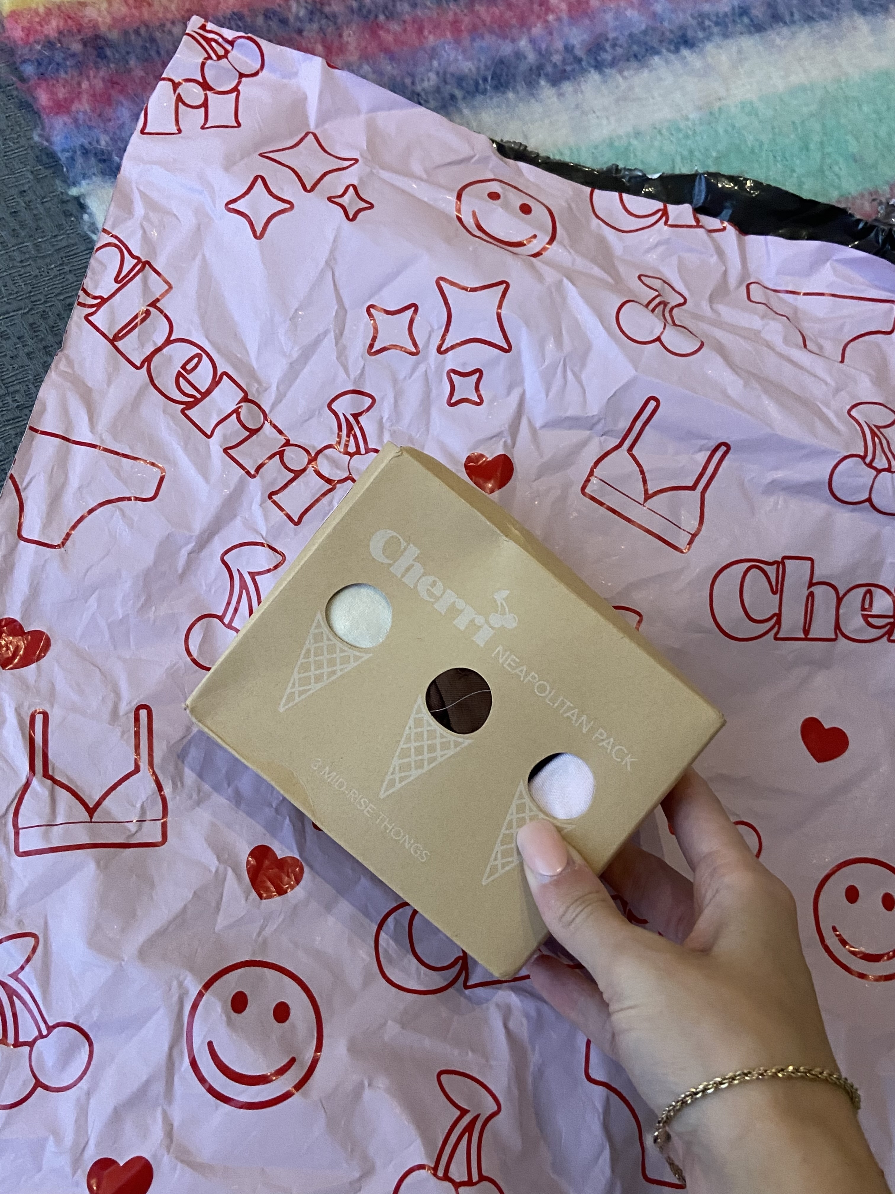 A hand holding a Cherri Neapolitan Pack box, placed on colorful Cherri-branded wrapping paper decorated with smiley faces, bras, stars, and hearts