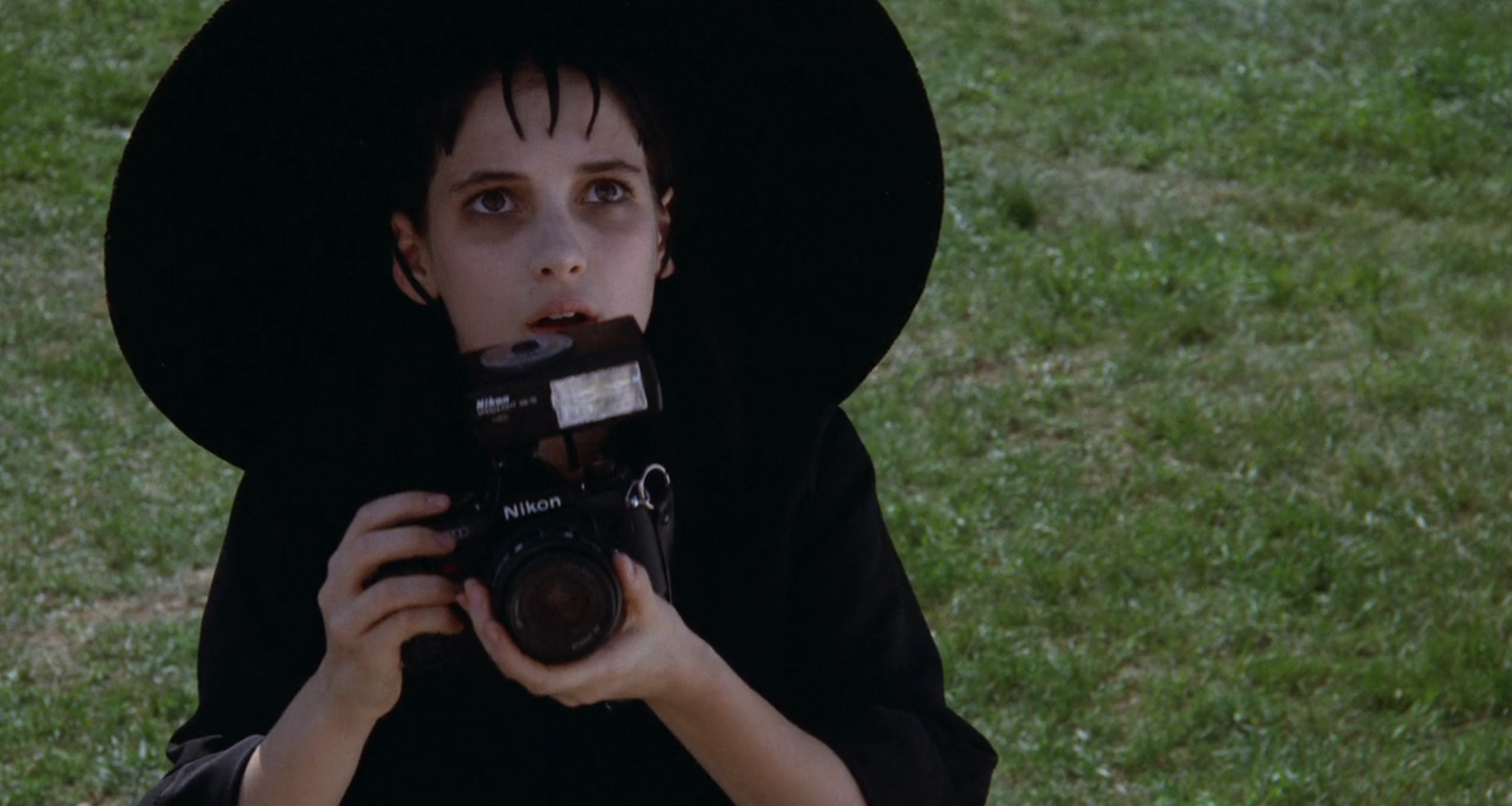 Winona Ryder in a wide-brimmed hat holds a camera, looking upward. She has a solemn expression and pale makeup