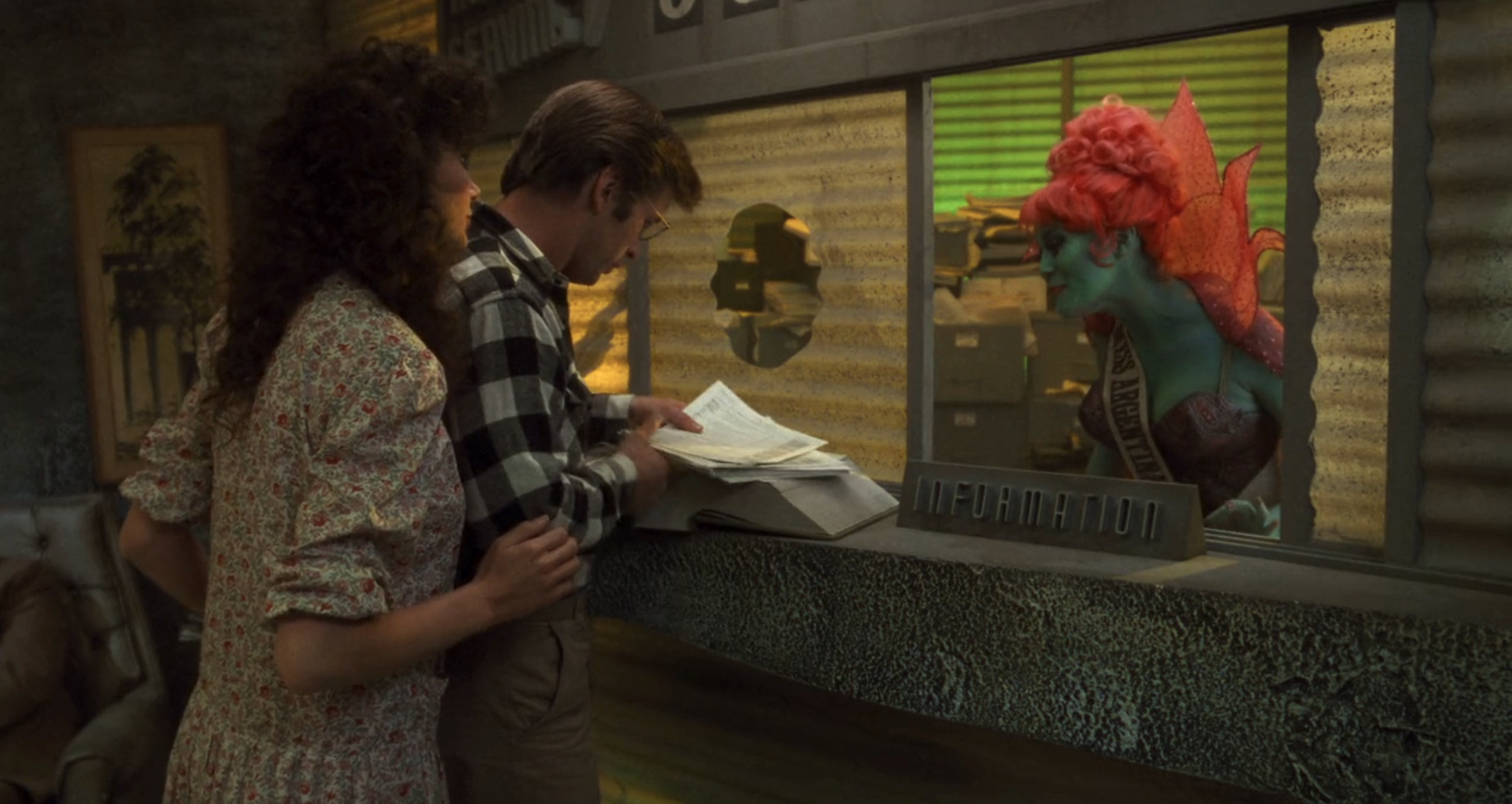 Geena Davis and Alec Baldwin speak to a green-skinned, Miss Argentina with fins at an information desk in a futuristic setting from the movie "Earth Girls Are Easy."