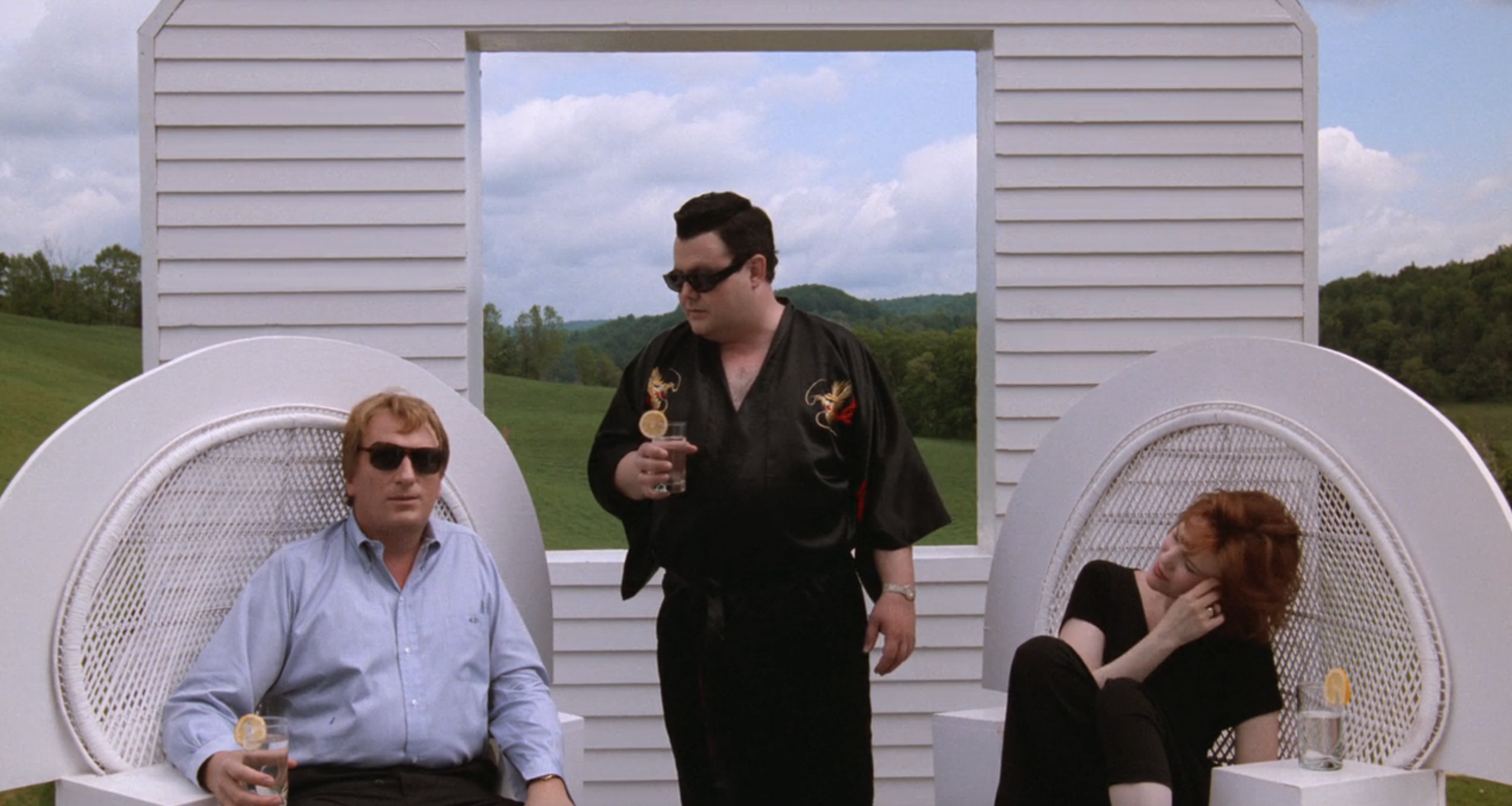 Jeffery Jones, dressed in a light shirt and dark sunglasses, poses with Glenn Shadix and Catherine O'Hara, who are also wearing sunglasses, seated on white chairs outdoors