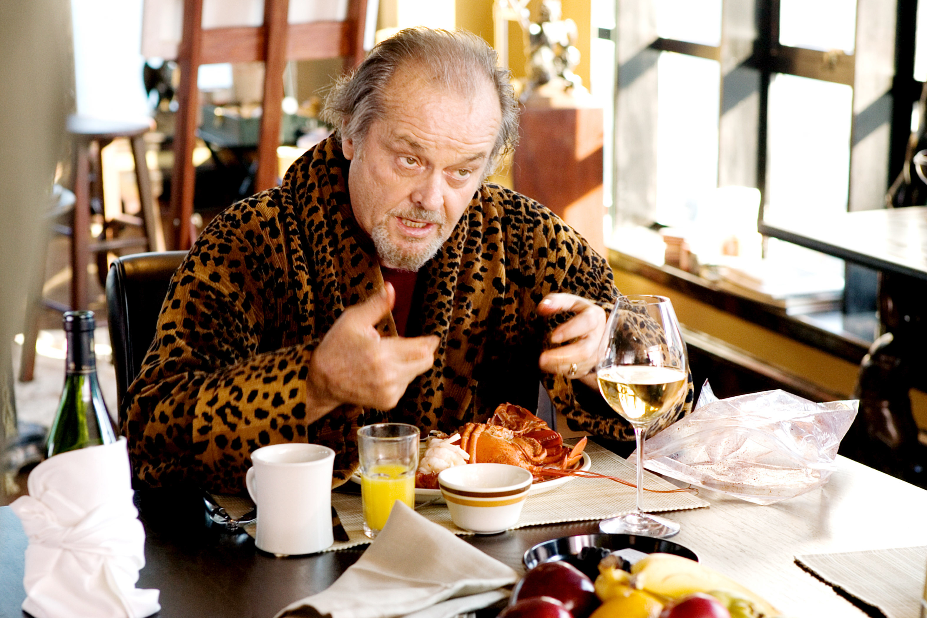 Jack Nicholson sits at a table in a leopard-print robe, gesturing with his hands while eating a meal, with a glass of wine and juice nearby
