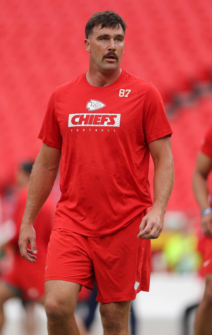 Travis Kelce wears an athletic Chiefs football shirt and shorts, standing on a field