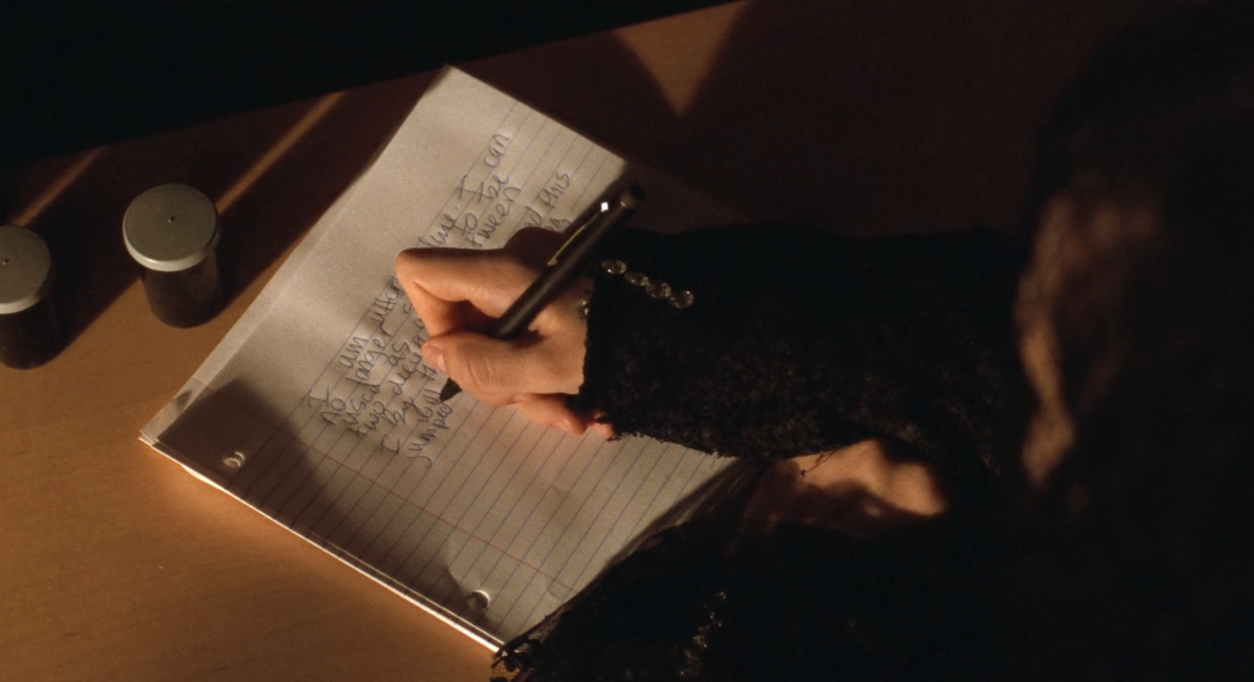 Lydia Deetz writes on a notebook with a pen. Text is barely legible. Two small containers are on the desk