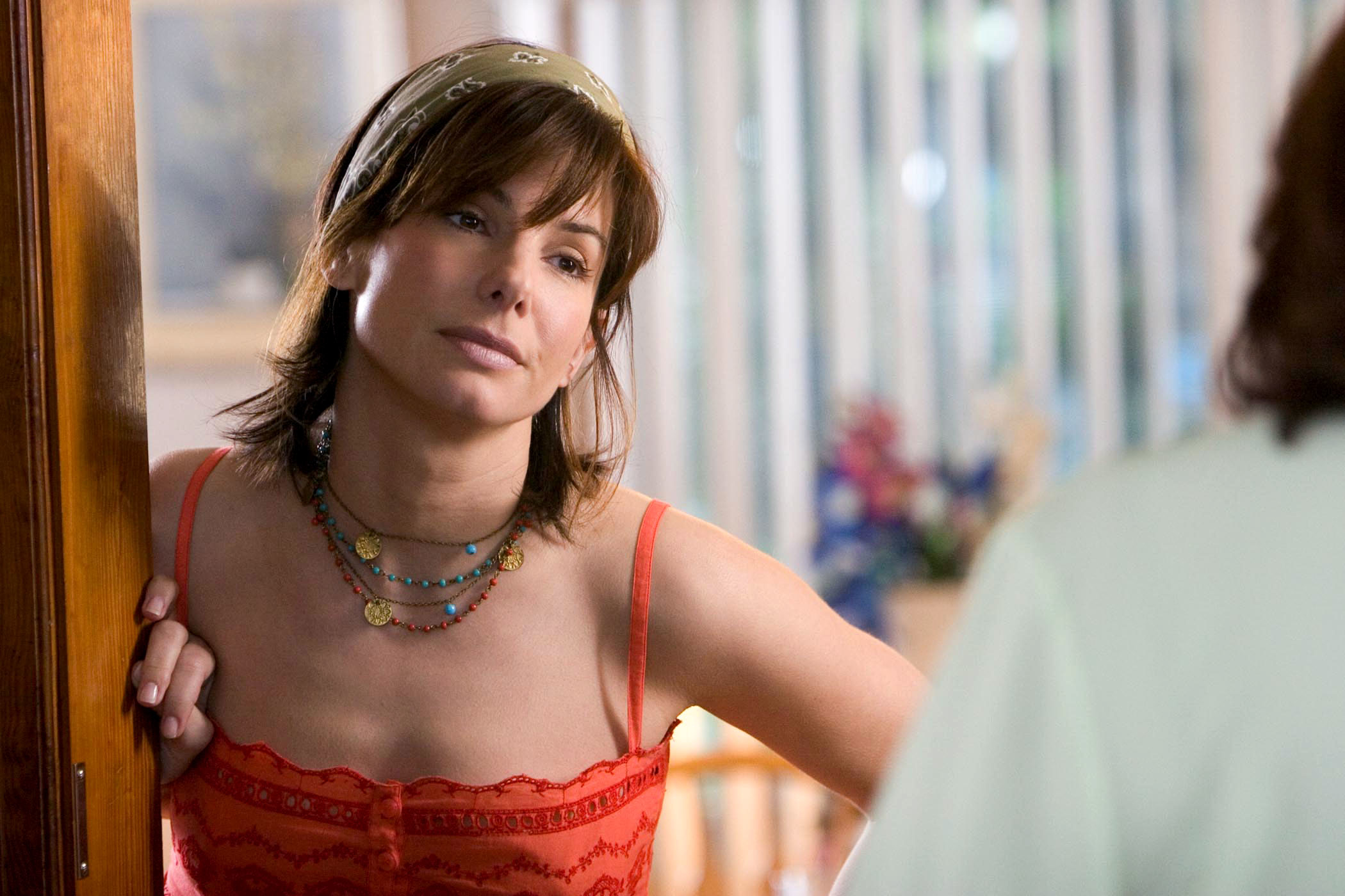 Sandra Bullock leaning against a door frame, wearing a casual sleeveless top and multi-strand necklaces, looking intently at someone off-camera