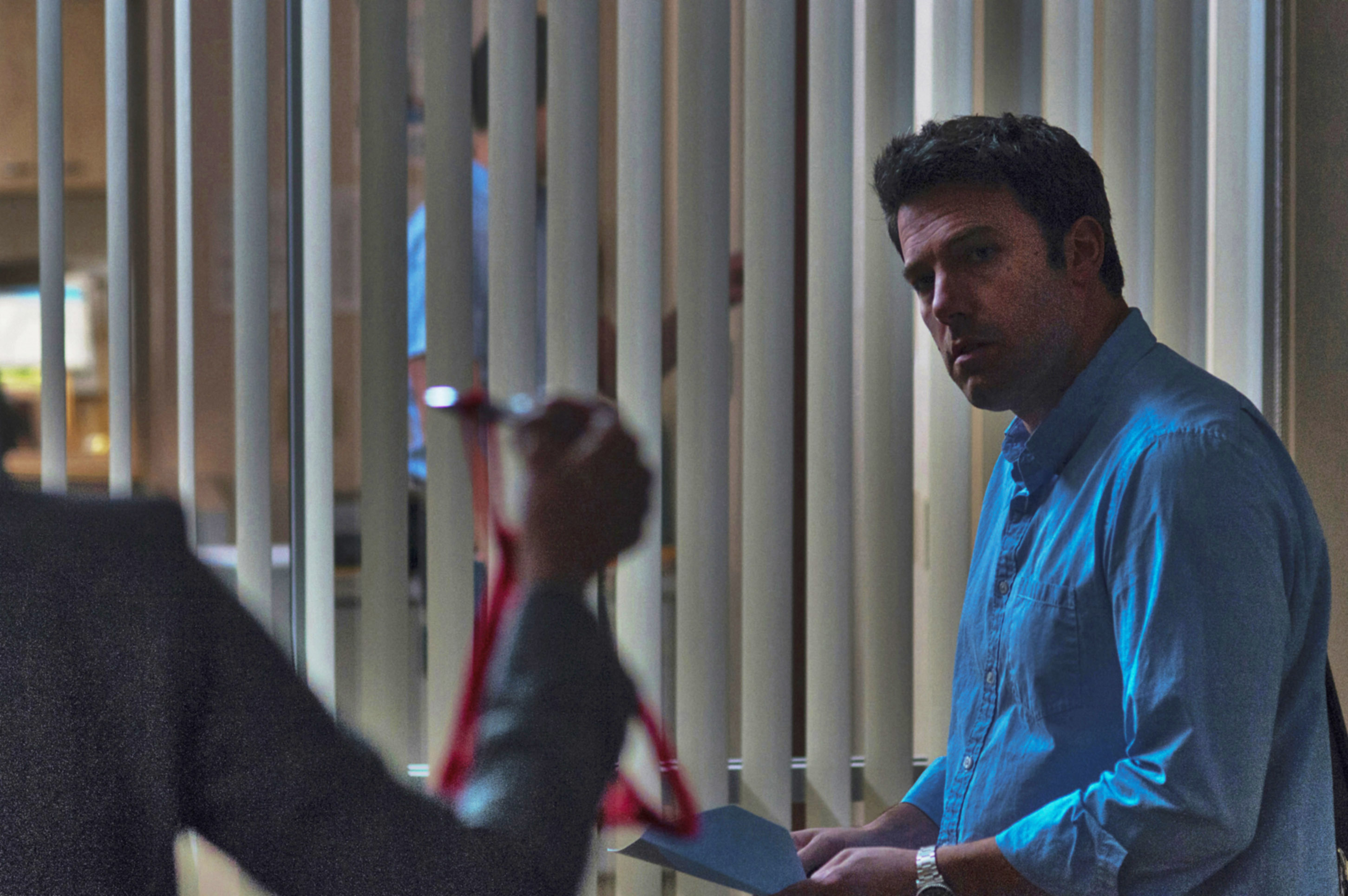 Ben Affleck stands indoors, holding paper, looking intently at someone out of frame who appears to be holding a lanyard
