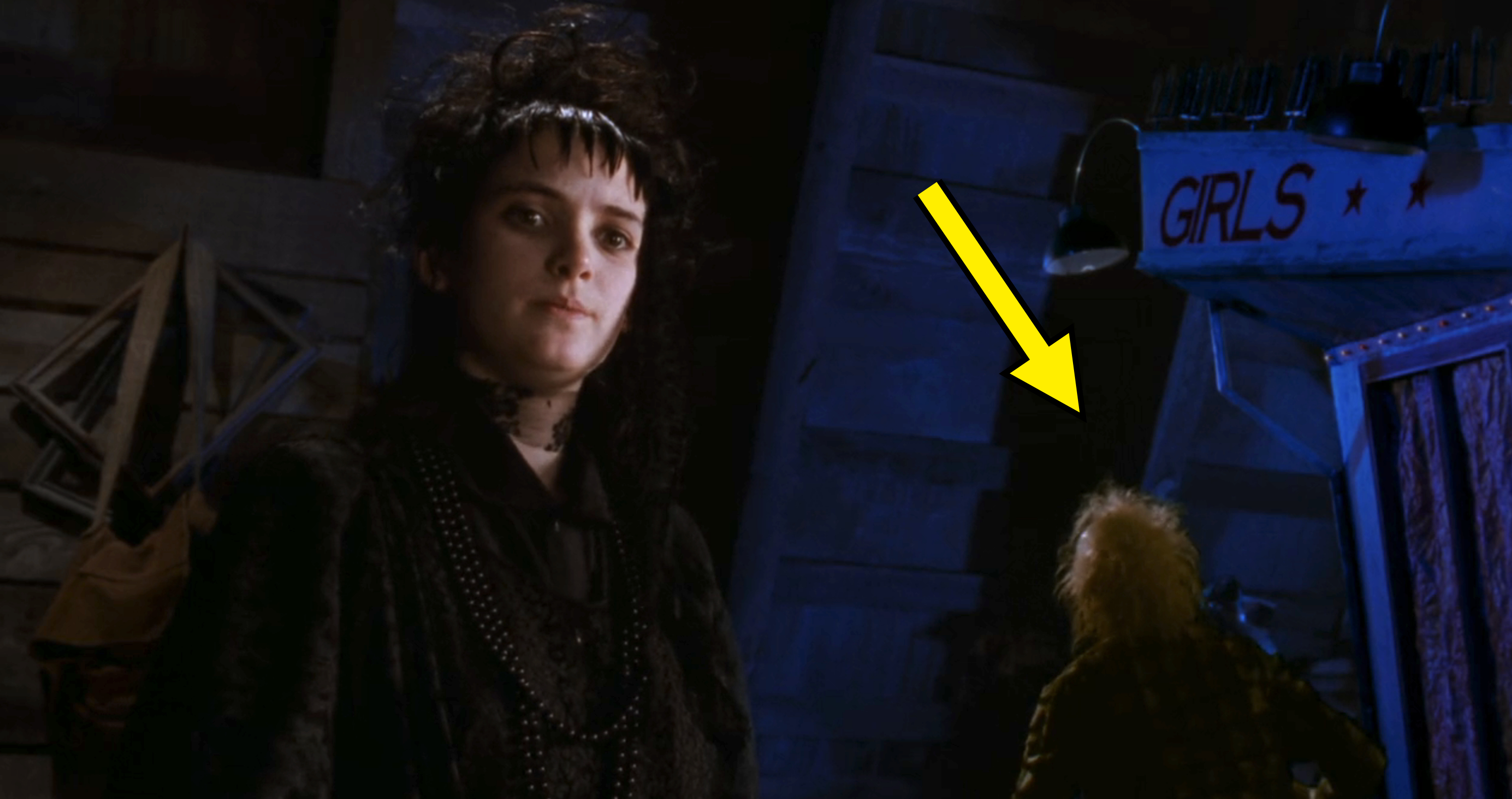Winona Ryder as Lydia Deetz in a gothic outfit, standing next to a figure near a sign reading "GIRLS" in a scene from the movie "Beetlejuice."