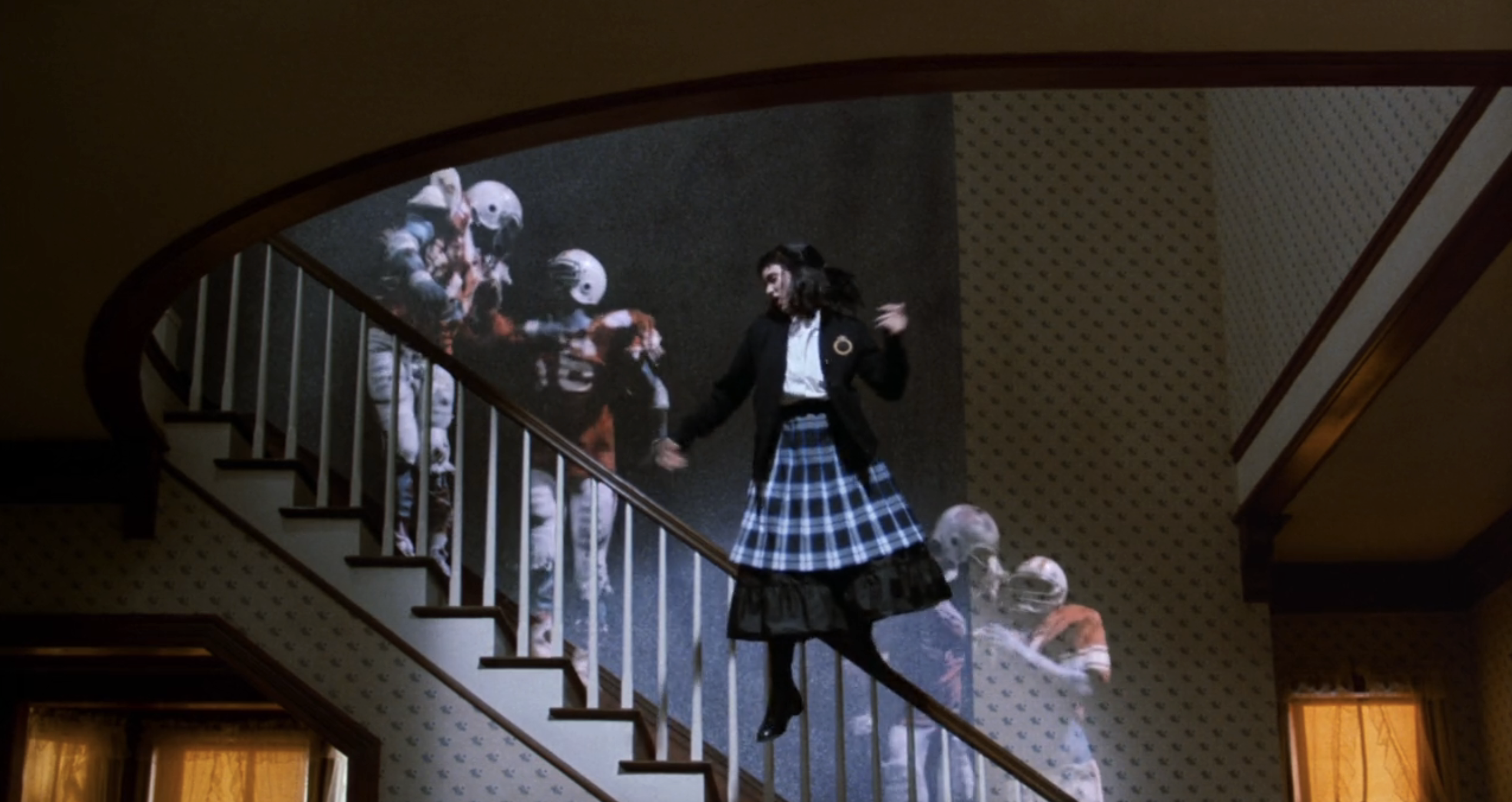 Winona Ryder stands on stairs, dressed in a dark outfit with a blue plaid skirt, with ghostly football players in the background