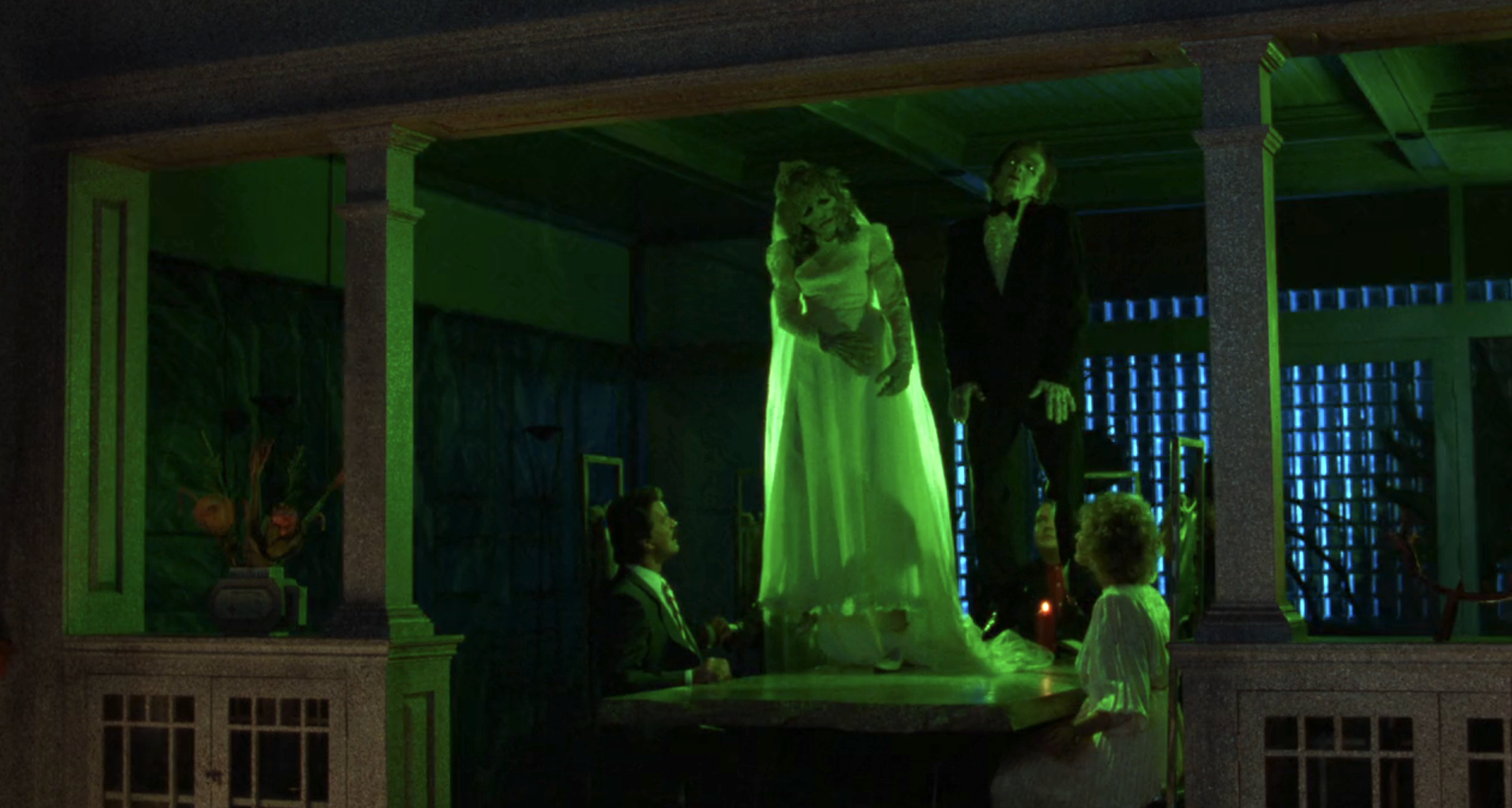 Two ghostly figures in formal attire float eerily above a table in a haunted-looking room. The scene is dimly lit and has a spooky ambiance from "Beetlejuice"
