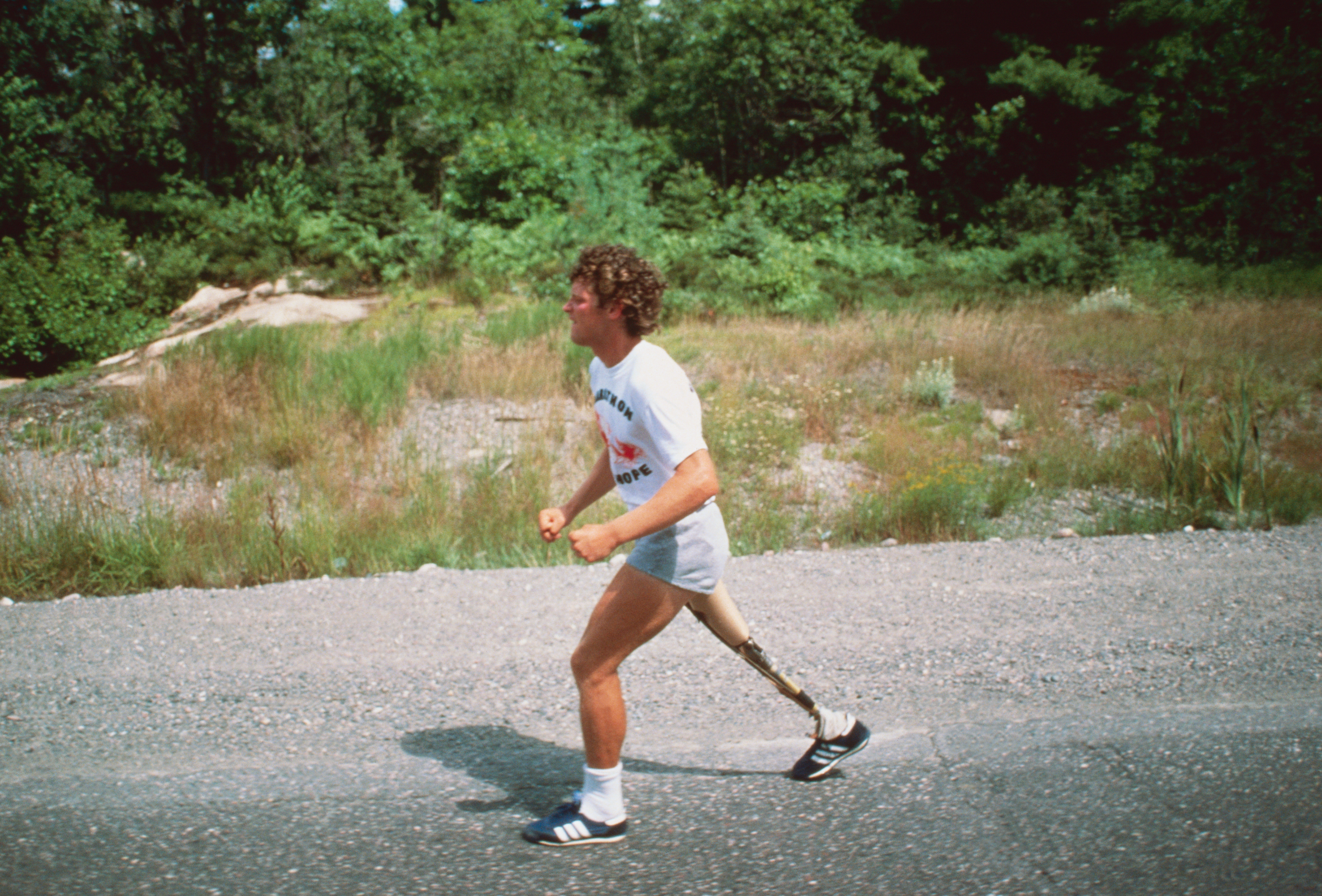 Terry Fox running on a road wearing a white T-shirt, shorts, and a prosthetic leg, with a forested background