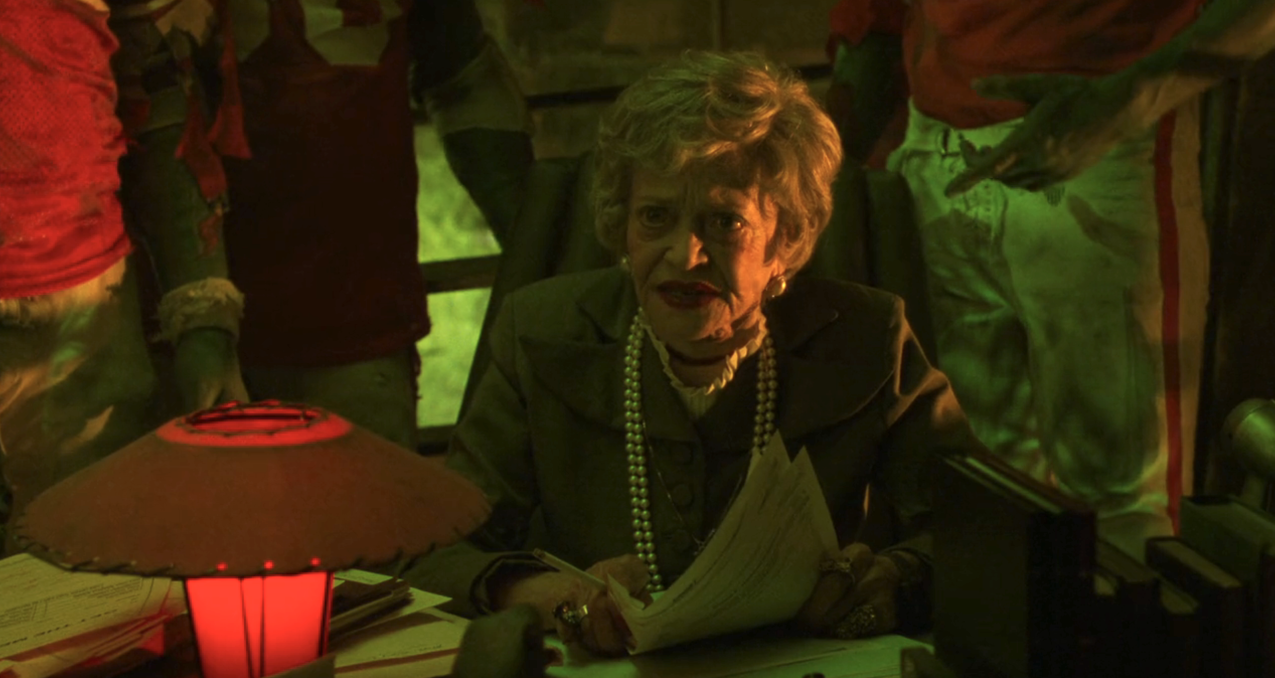 Sylvia Sidney with a formal outfit and pearl necklace sits at a desk holding papers, surrounded by people in football uniforms
