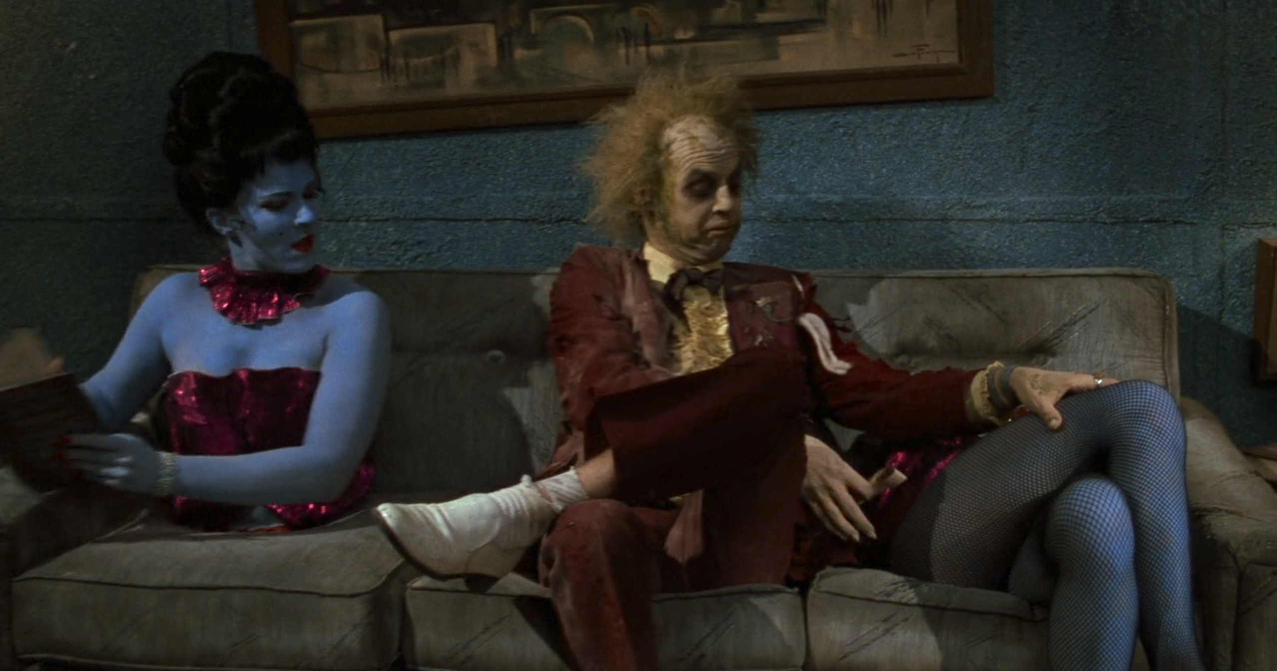 Two characters: a blue-skinned person in a red sequined outfit seated next to a disheveled Michael Keaton with wild hair wearing a torn suit and fishnet stockings