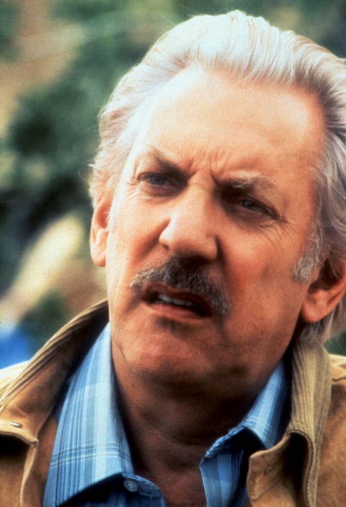Donald Sutherland in a scene from a film. He has a serious expression and is wearing a striped shirt and jacket