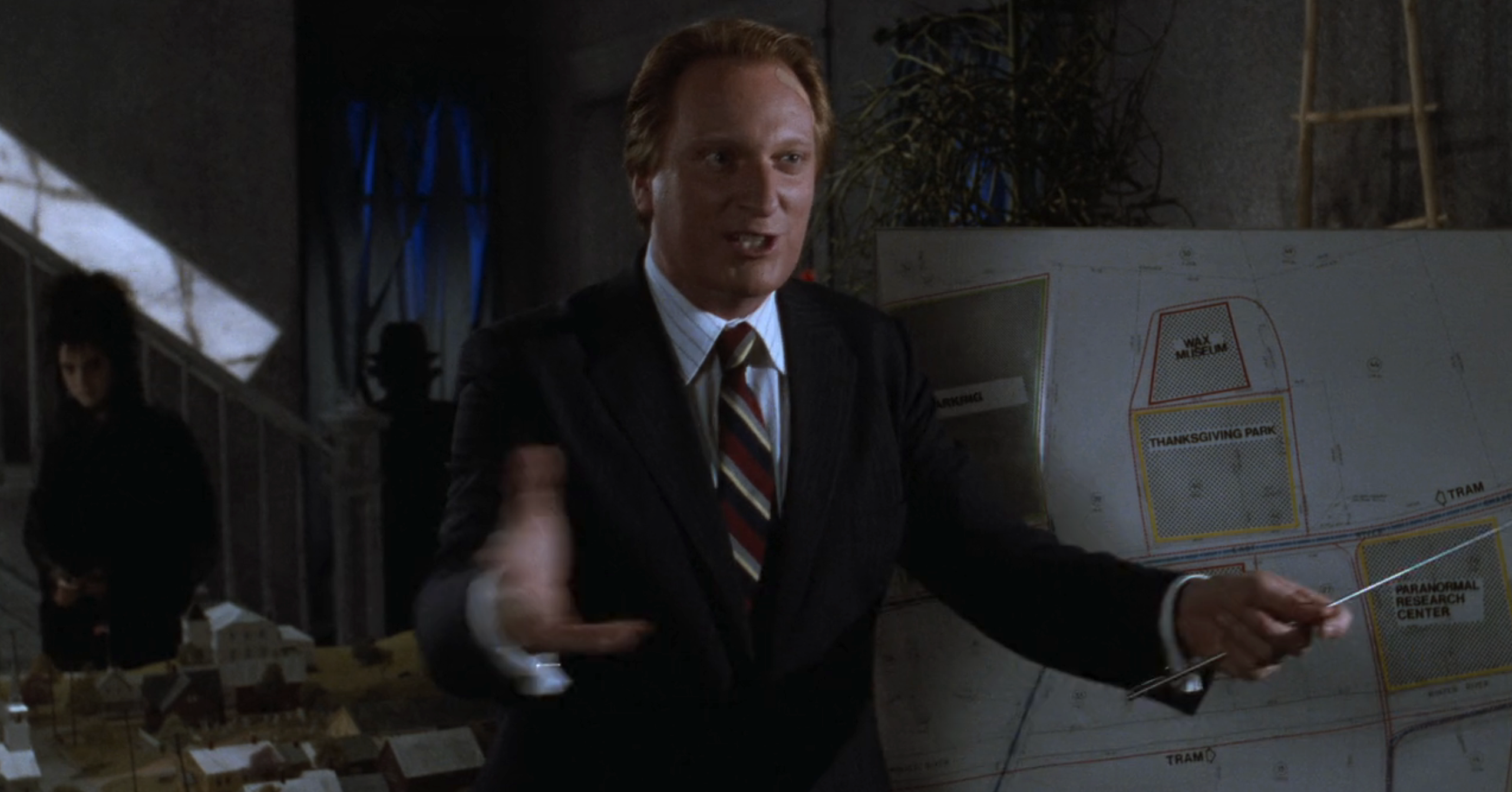 Jeffery Jones in a suit and striped tie gestures while standing next to a model of a town and a large map in an interior setting
