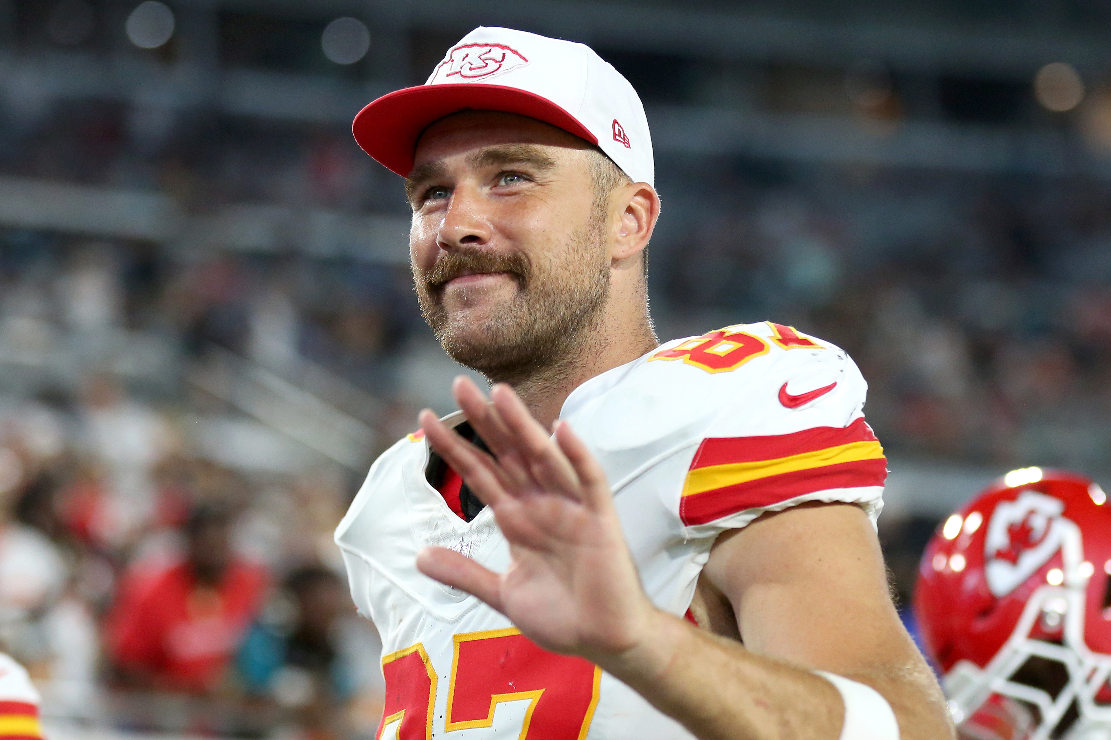 Travis Kelce waves while wearing a white Kansas City Chiefs uniform and a matching cap at a sporting event