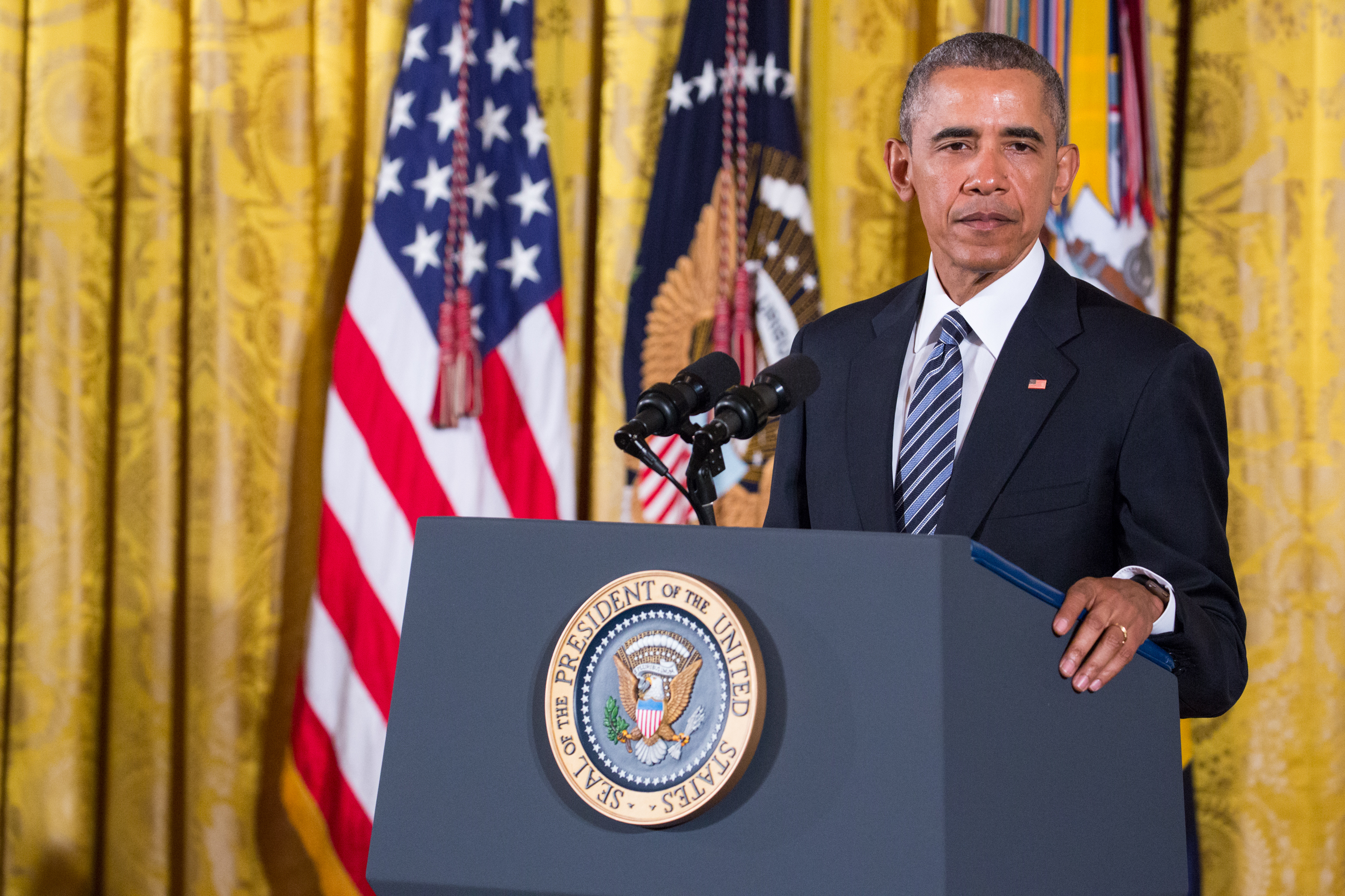 Barack Obama stands at a podium with the U.S. presidential seal, speaking during an event, with American flags in the background