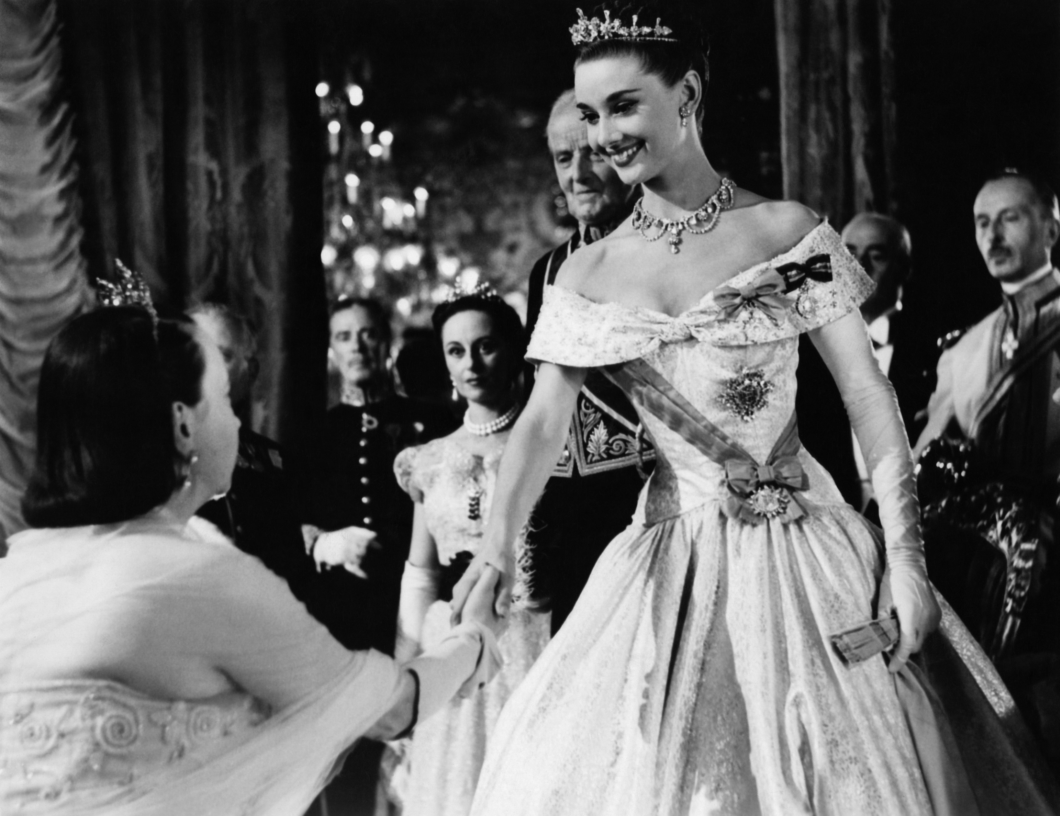 Audrey Hepburn, in an elegant ball gown and tiara, shakes hands with another woman in formal attire during a grand event with others looking on