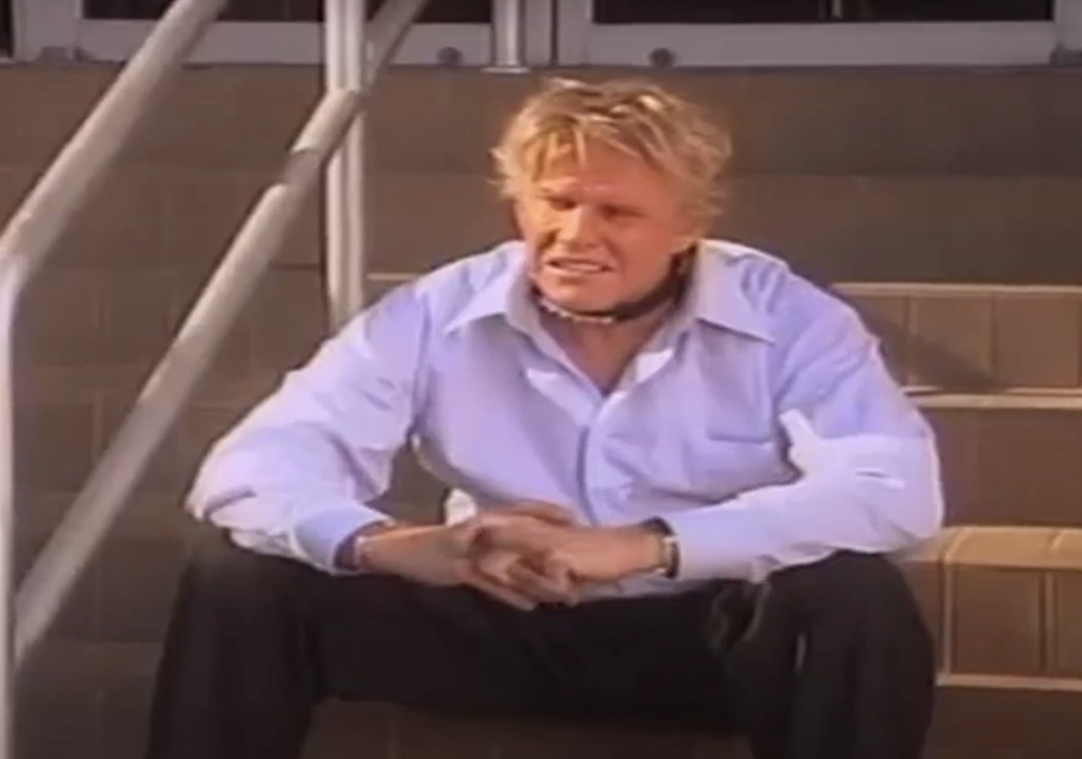 Gary Busey with blond hair, wearing a collared shirt and dark pants, sits on stairs