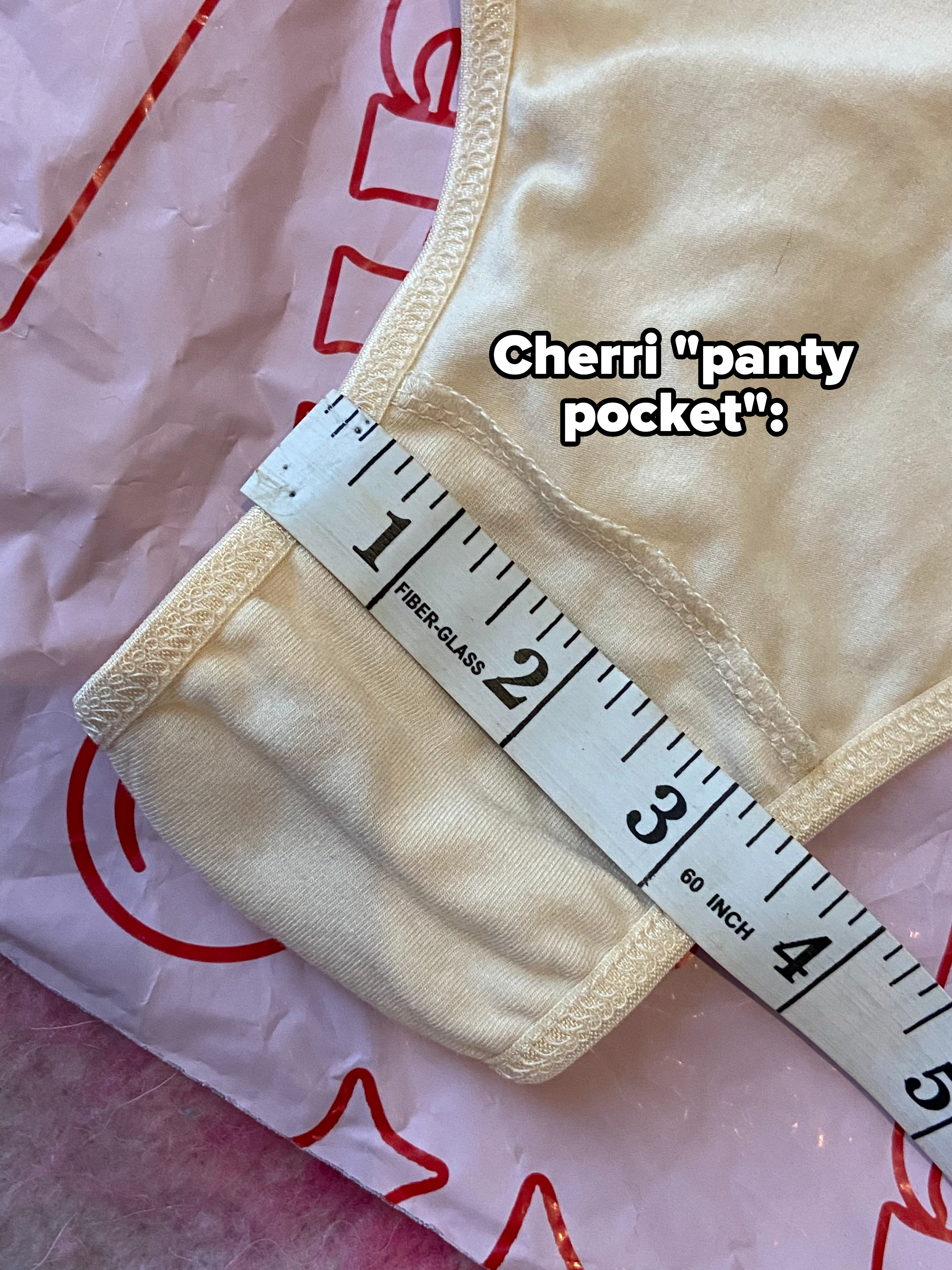 A measuring tape is laid across a fabric item, showing a measurement of about 2 inches on the fabric. An indistinct bag is partially visible beneath the fabric