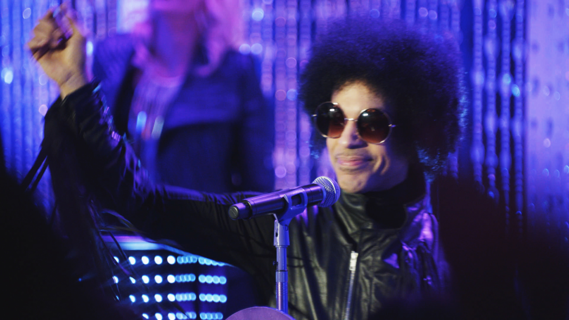 Prince on stage, wearing sunglasses and a leather jacket, performing energetically with a microphone in hand