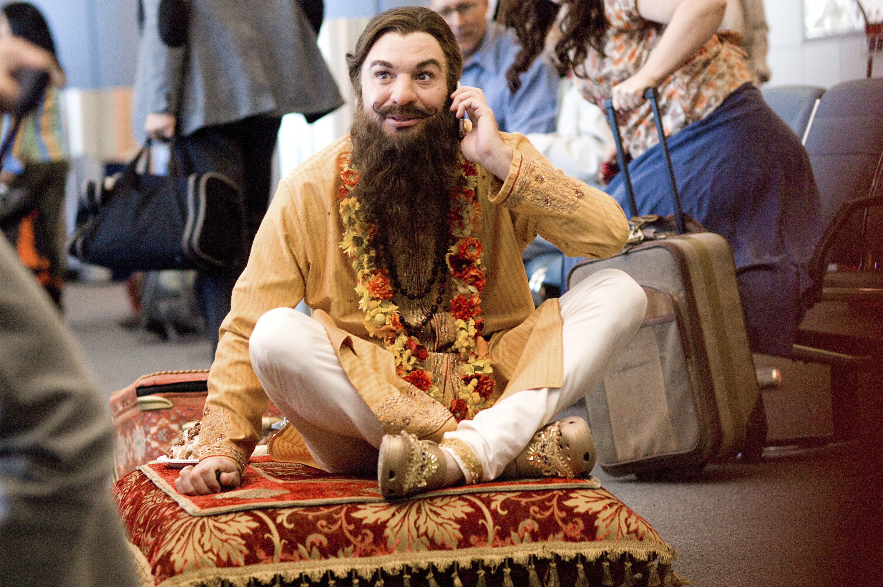 Mike Myers with a long beard, wearing traditional Indian attire with floral garlands, sitting cross-legged on a patterned rug, talking on a phone at an airport