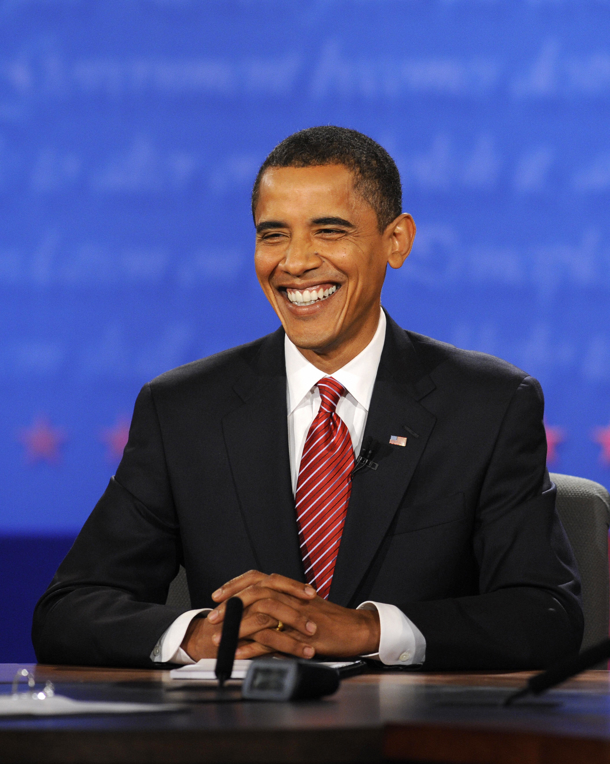 Barack Obama sits at a desk, smiling and wearing a suit with a striped tie. He appears relaxed and confident