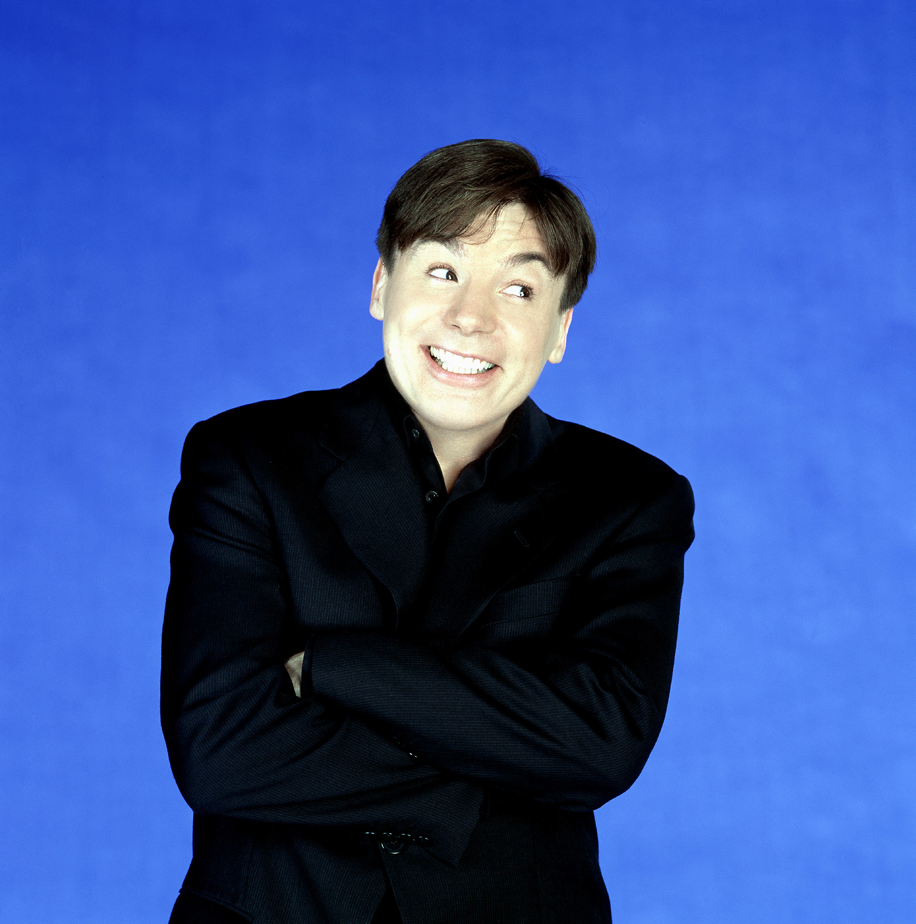 Mike Myers smiling with arms crossed, wearing a dark suit against a plain background