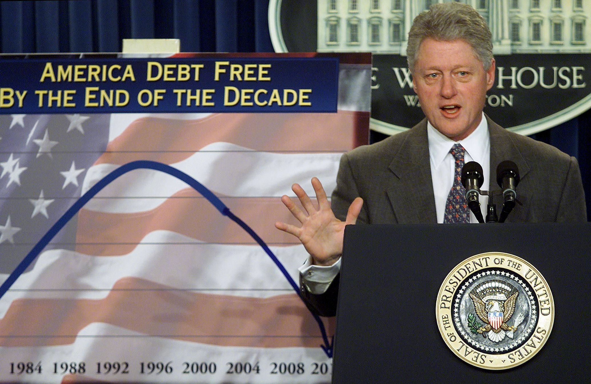Bill Clinton speaks at a podium with a chart showing national debt trends in the background. Text on the chart reads "America Debt Free By The End Of The Decade."