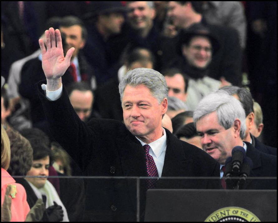 Bill Clinton waves to a crowd from behind a podium while Newt Gingrich stands nearby, surrounded by other individuals