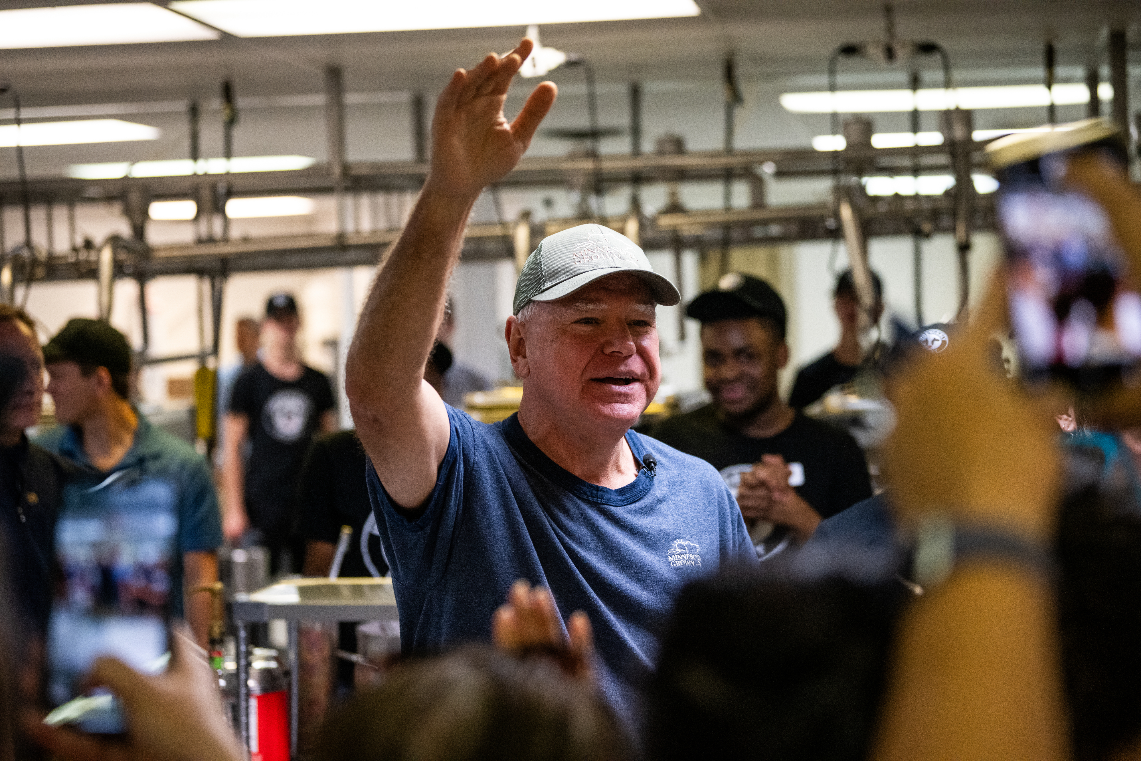 A man in a blue shirt greets a crowd in what appears to be a factory or workshop setting. Several people in the background observe and take photos