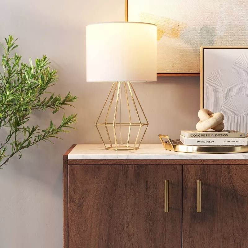 A modern table lamp with a geometric base sits on a wooden cabinet alongside a potted plant, framed artwork, and a stack of design books