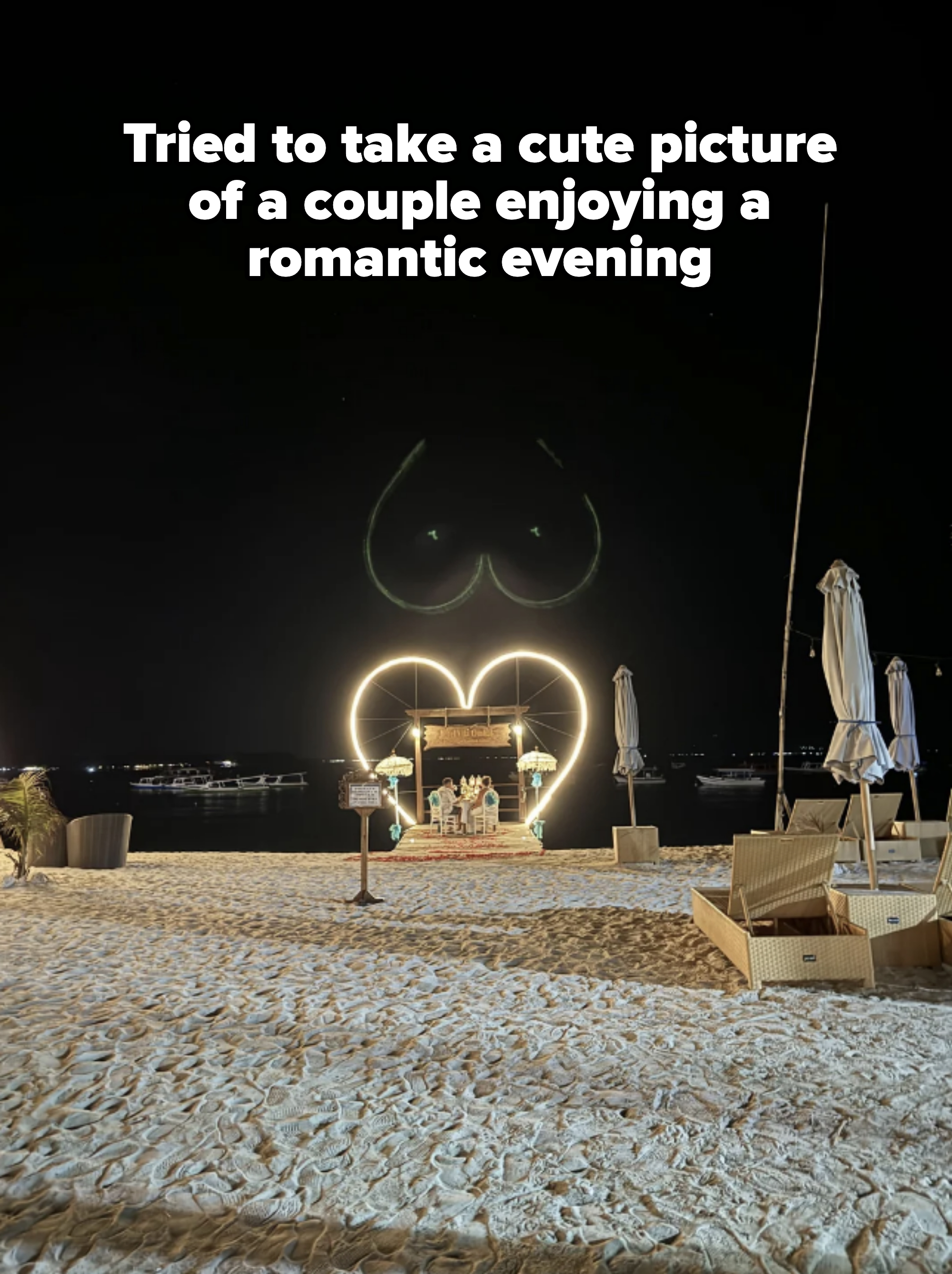Heart-shaped light decoration on a beach at night with lounge chairs and umbrellas nearby. No people are visible