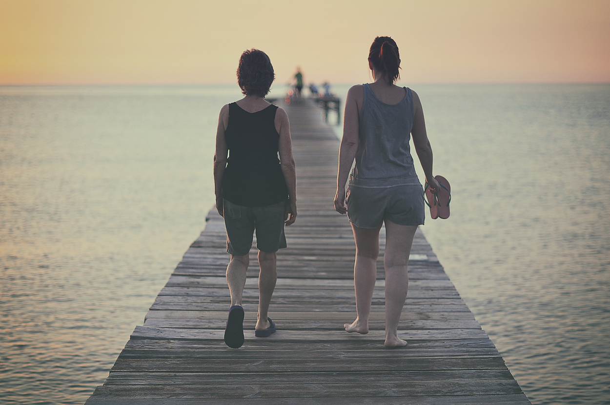 Two people, seen from behind, walking barefoot on a wooden pier towards the horizon. The person on the right holds sandals in their hand