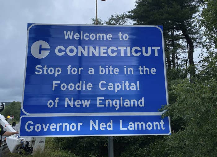 Welcome to Connecticut sign reads &quot;Stop for a bite in the Foodie Capital of New England&quot; with &quot;Governor Ned Lamont&quot; at the bottom. Forest background