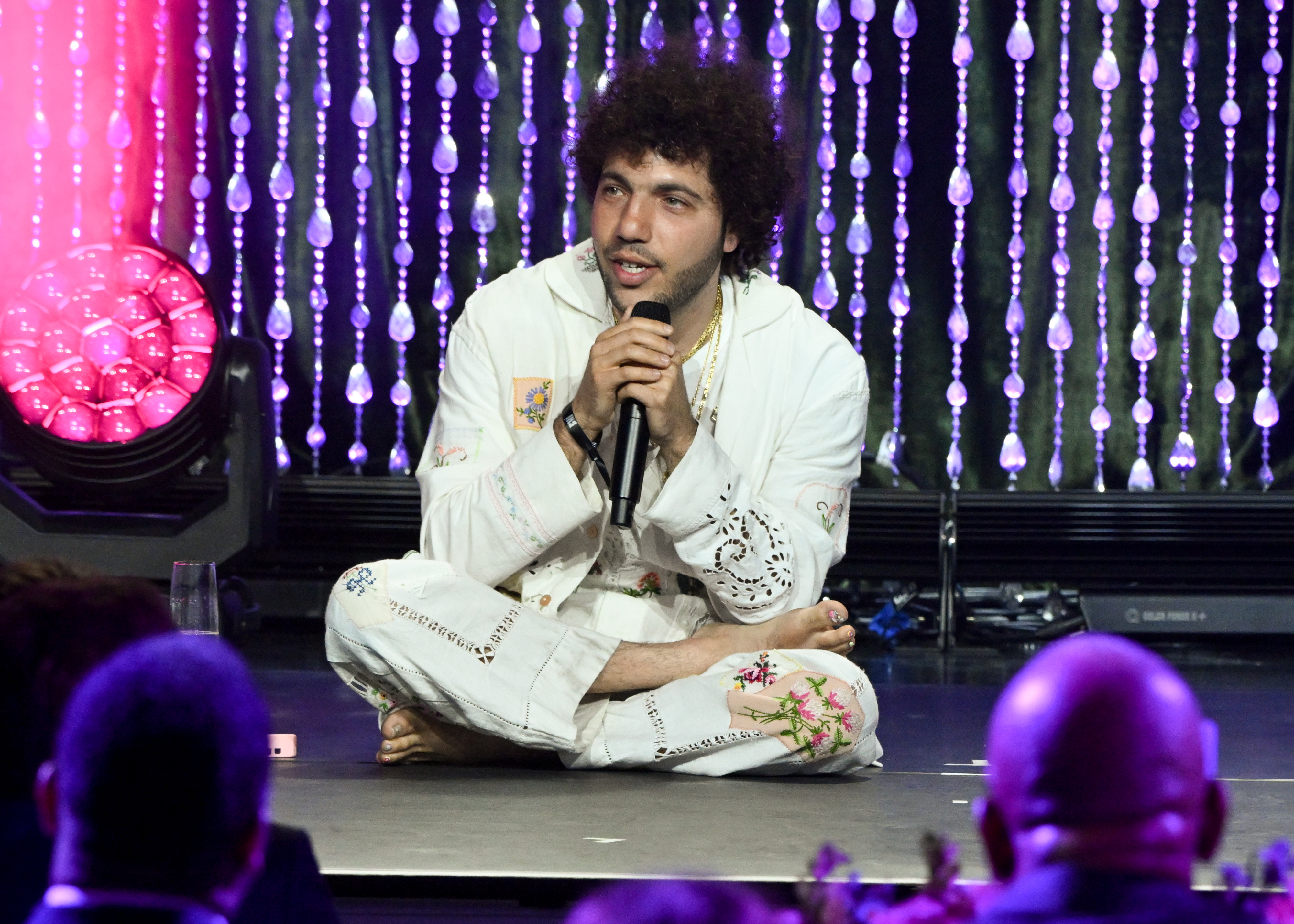 Benny Blanco sits cross-legged on stage, wearing white embroidered attire, holding a microphone, with a backdrop of hanging lights