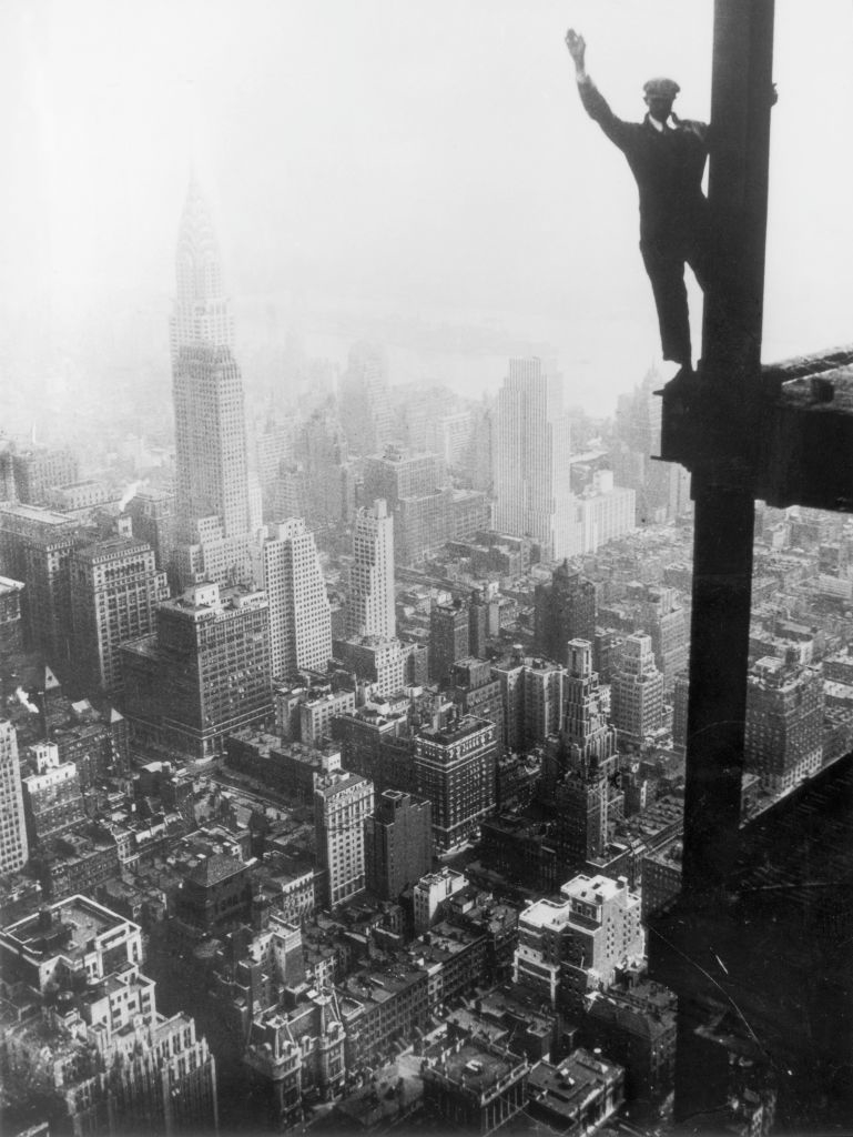 Man standing on a steel beam high above New York City, likely during the construction of a skyscraper. His right hand is raised in a wave