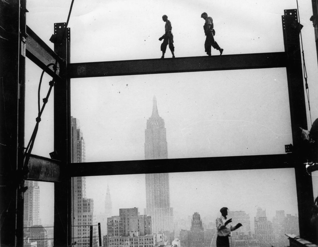 Two construction workers walk on a steel beam high above New York City, with the Empire State Building visible in the background. Another worker stands below