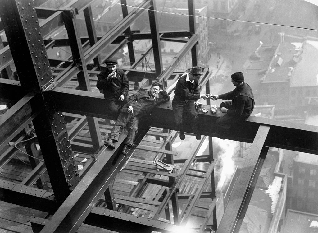 Four construction workers sit on a steel beam high above the ground, taking a break; they appear relaxed and possibly eating or talking