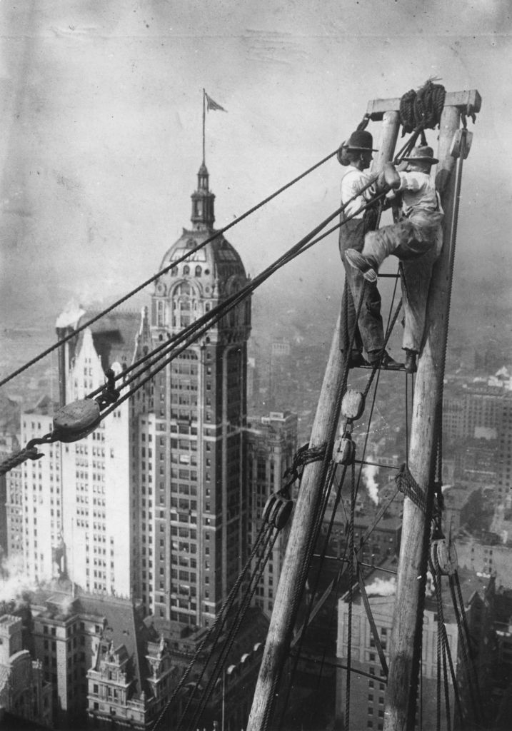 Two workers are perched on a tall wooden structure performing construction work high above a city skyline. The dense urban landscape is visible below