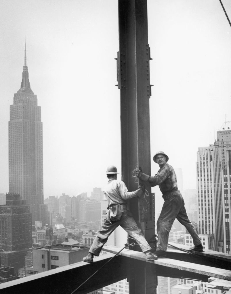 Two workers in overalls are balancing on steel beams high above New York City while constructing a building, with the Empire State Building in the background