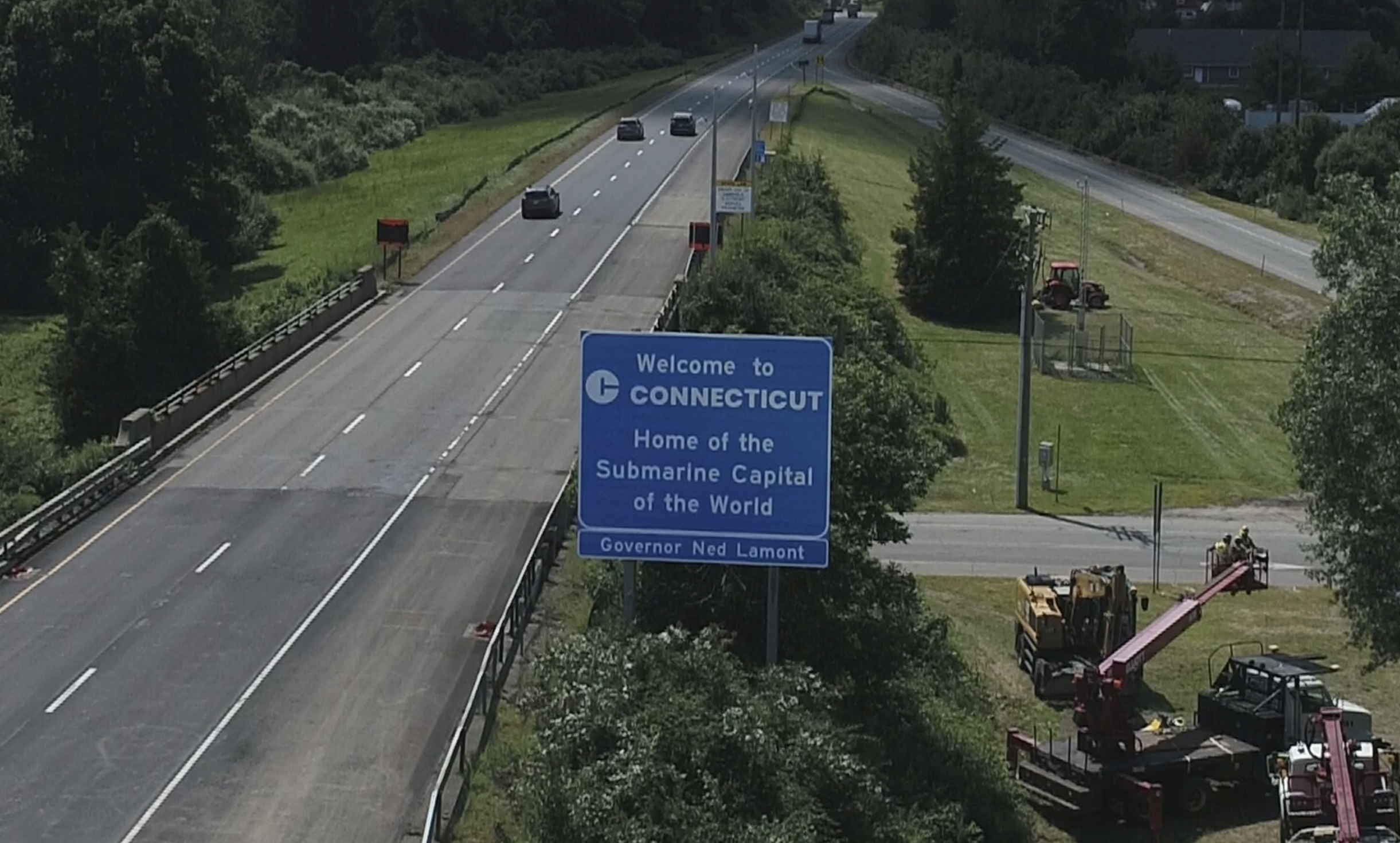 A highway sign reads &quot;Welcome to Connecticut, Home of the Submarine Capital of the World - Governor Ned Lamont.&quot; Machinery is visible on the roadside