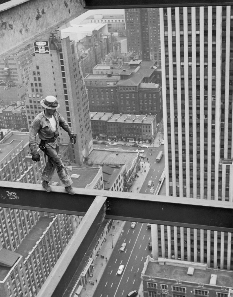 A construction worker balances on a steel beam high above the city streets amidst skyscrapers. The image captures the height and risk of the job