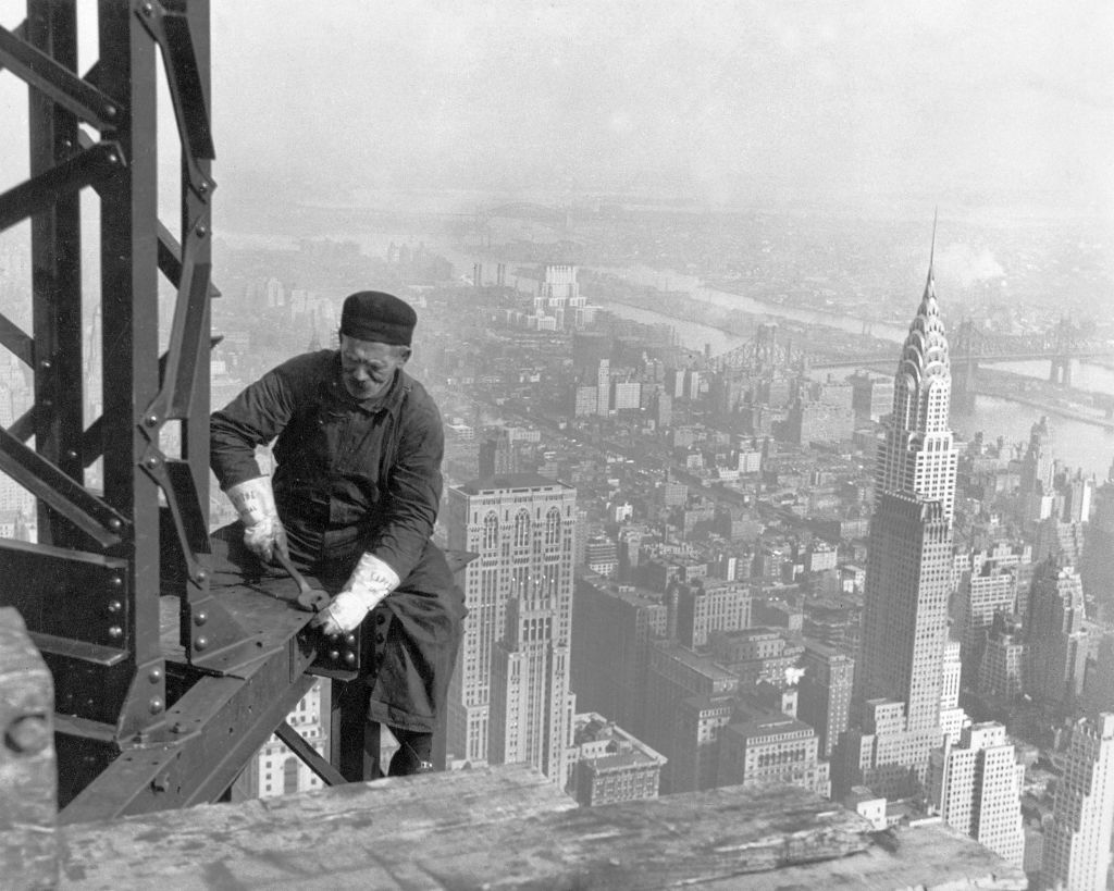 A worker in overalls and a cap is seen on an iron beam high above New York City during the construction of a skyscraper in the early 20th century