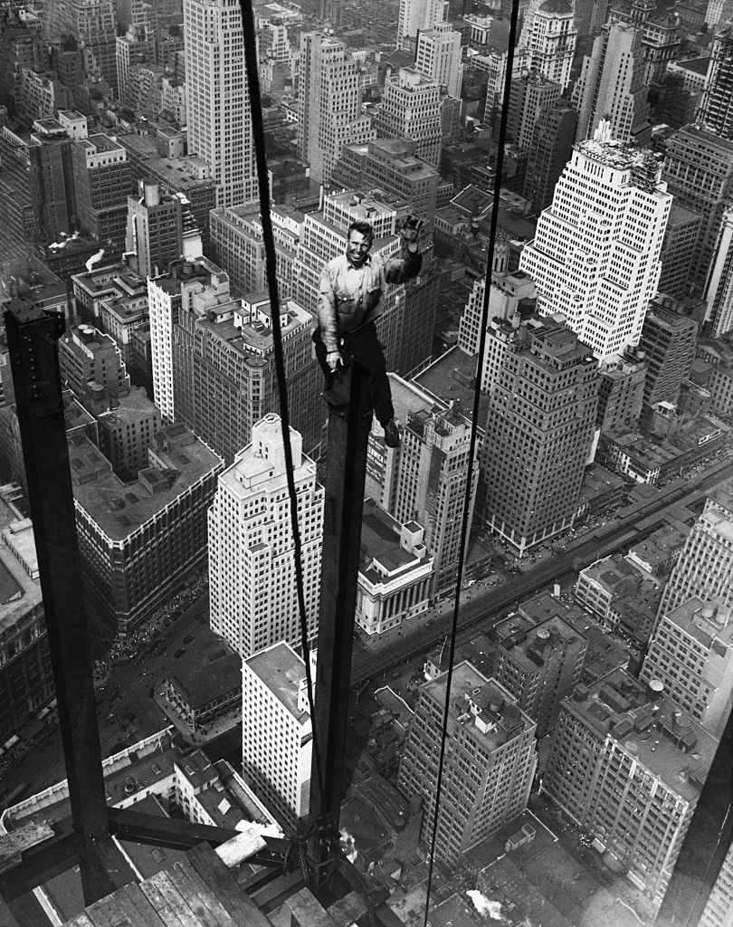 A construction worker stands on a steel beam high above a city skyline, waving at the camera. The image shows an old cityscape with tall buildings